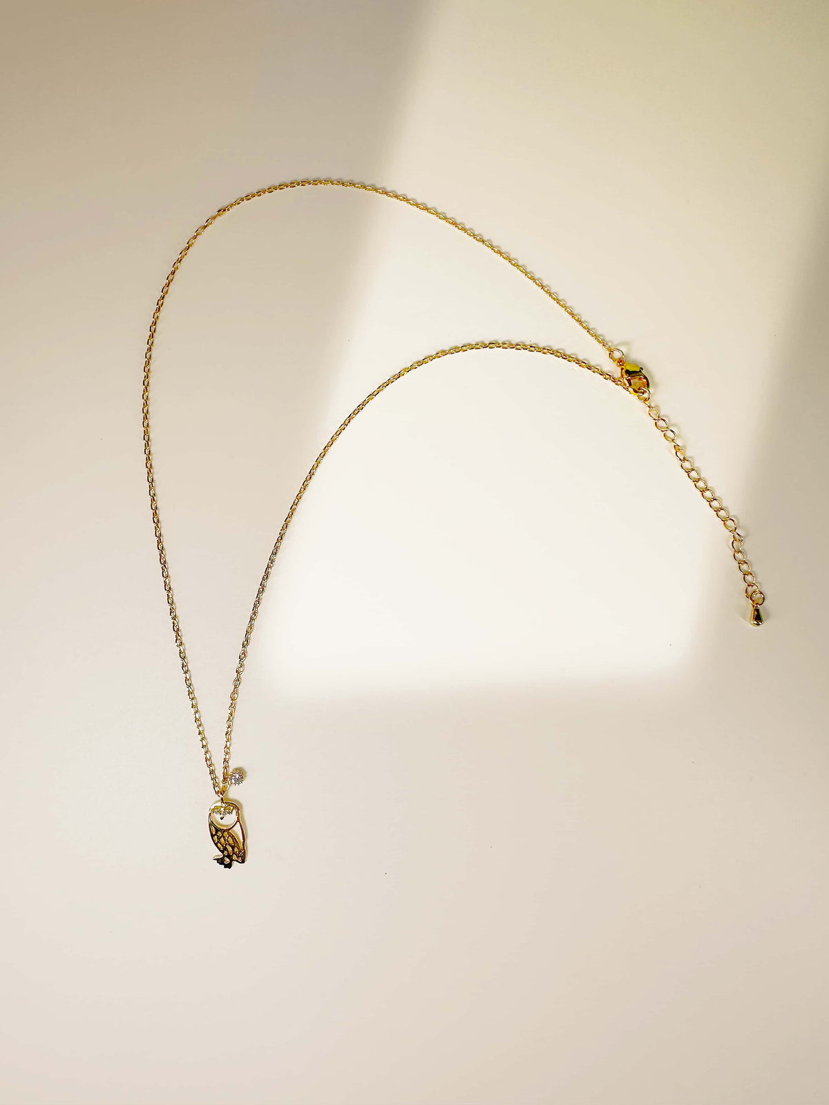 Full flat lay of the 18K gold owl necklace with an adjustable chain and lobster clasp on a minimalist warm backdrop