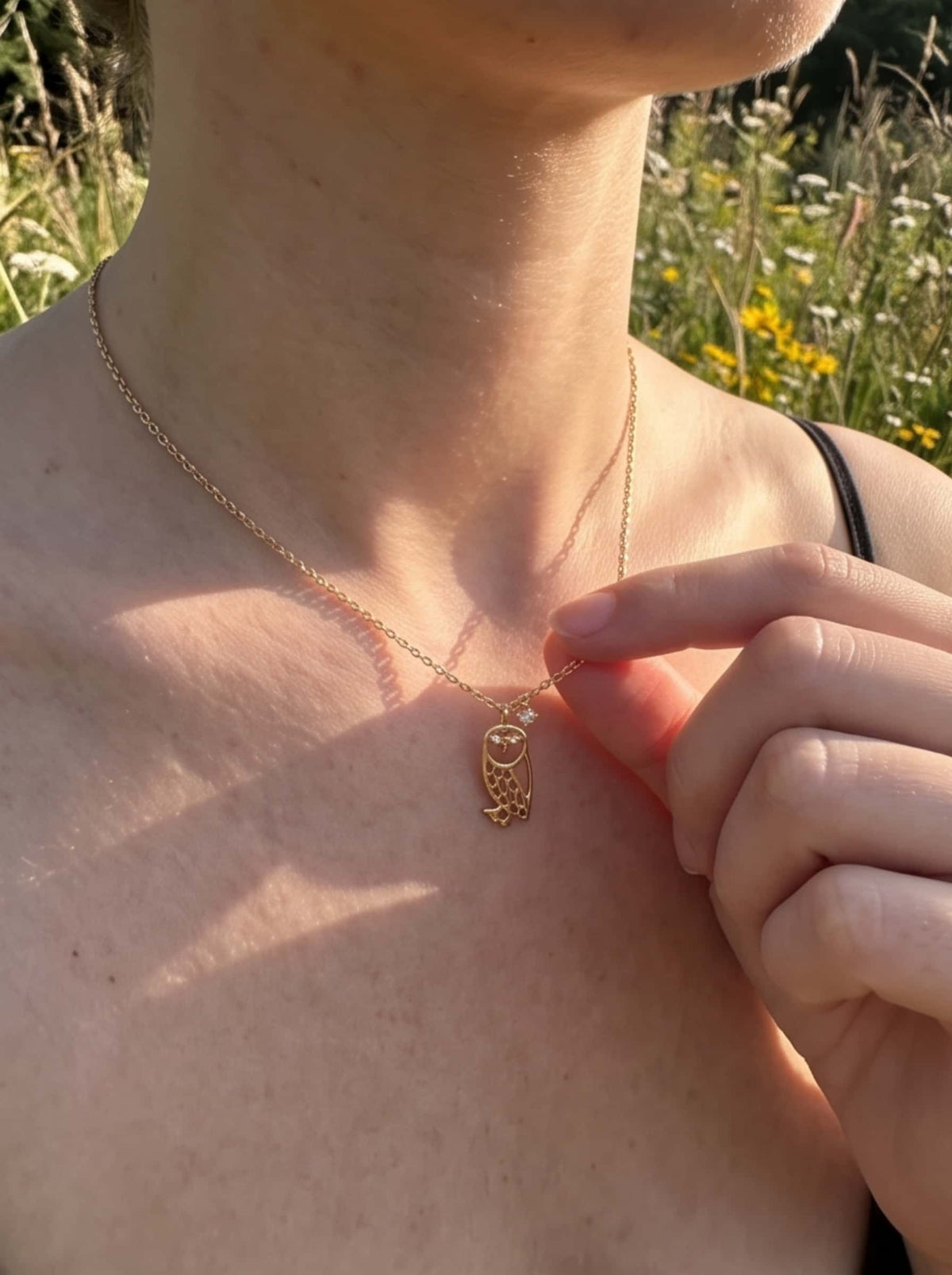 Close-up of a model gently holding the dainty 18K gold owl pendant outdoors in golden natural sunlight