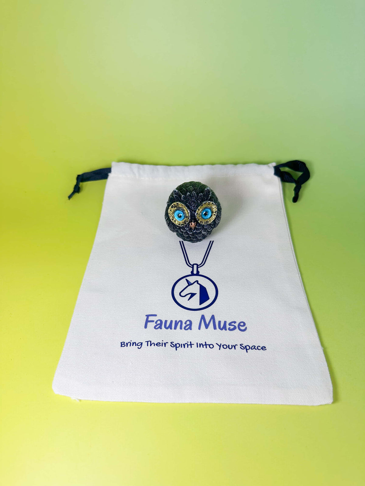 A small owl decor with blue and gold eyes on a black cord, displayed on top of a white drawstring bag printed with the Fauna Muse logo and text, set against a yellow-green gradient background.