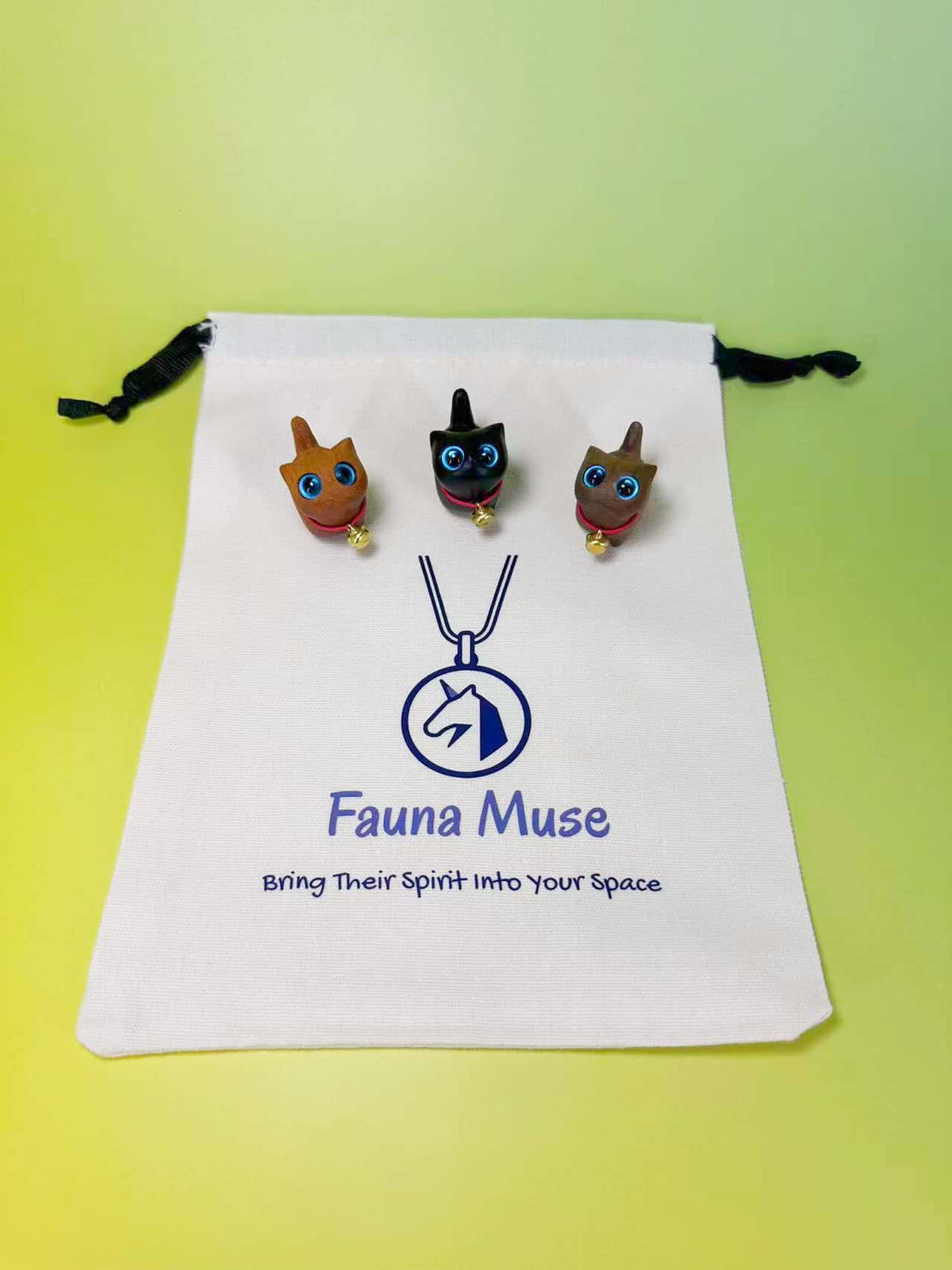 Three small cat figurines displayed on a white drawstring pouch printed with the Fauna Muse logo and slogan, set against a bright yellow-green gradient background.