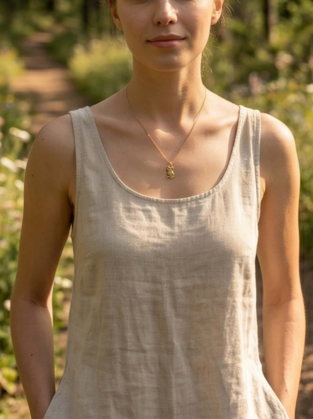 A woman in a neutral linen tank top wearing the delicate gold owl necklace while standing on a sunlit forest trail