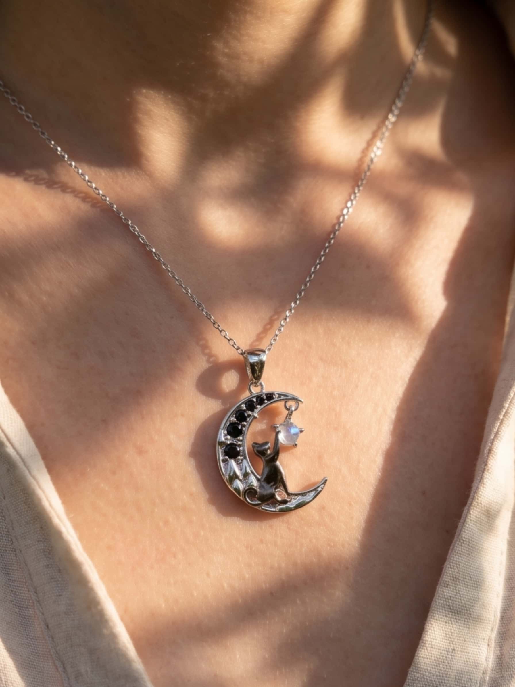 Close up of a beautifully crafted cat pendant necklace resting on a collarbone, featuring a black cat reaching for a shining moonstone