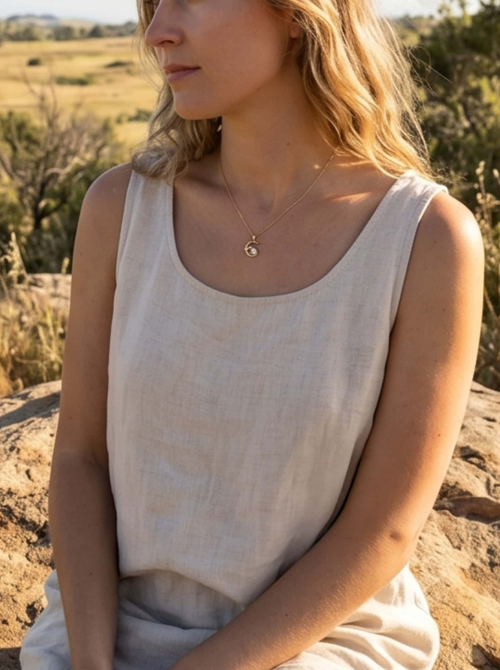 Lifestyle photography of a woman in a cottagecore linen outfit wearing a gold cat moon necklace, sitting peacefully on a rock in a sunny field