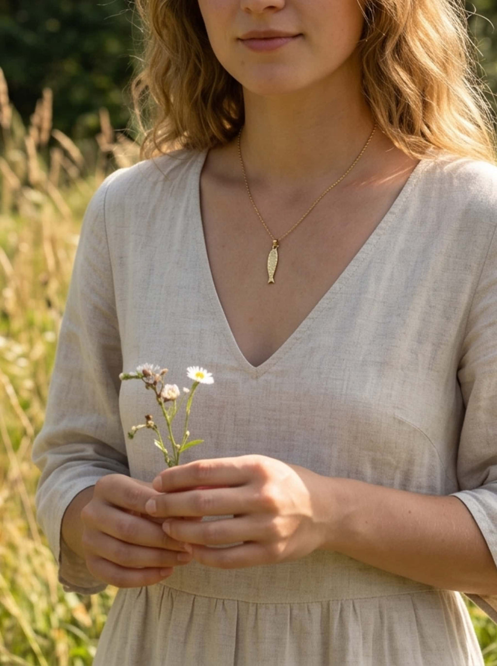 Close-up of a model in a cottagecore linen dress wearing a fish necklace while gently holding small white wild daisies in a sunny summer meadow