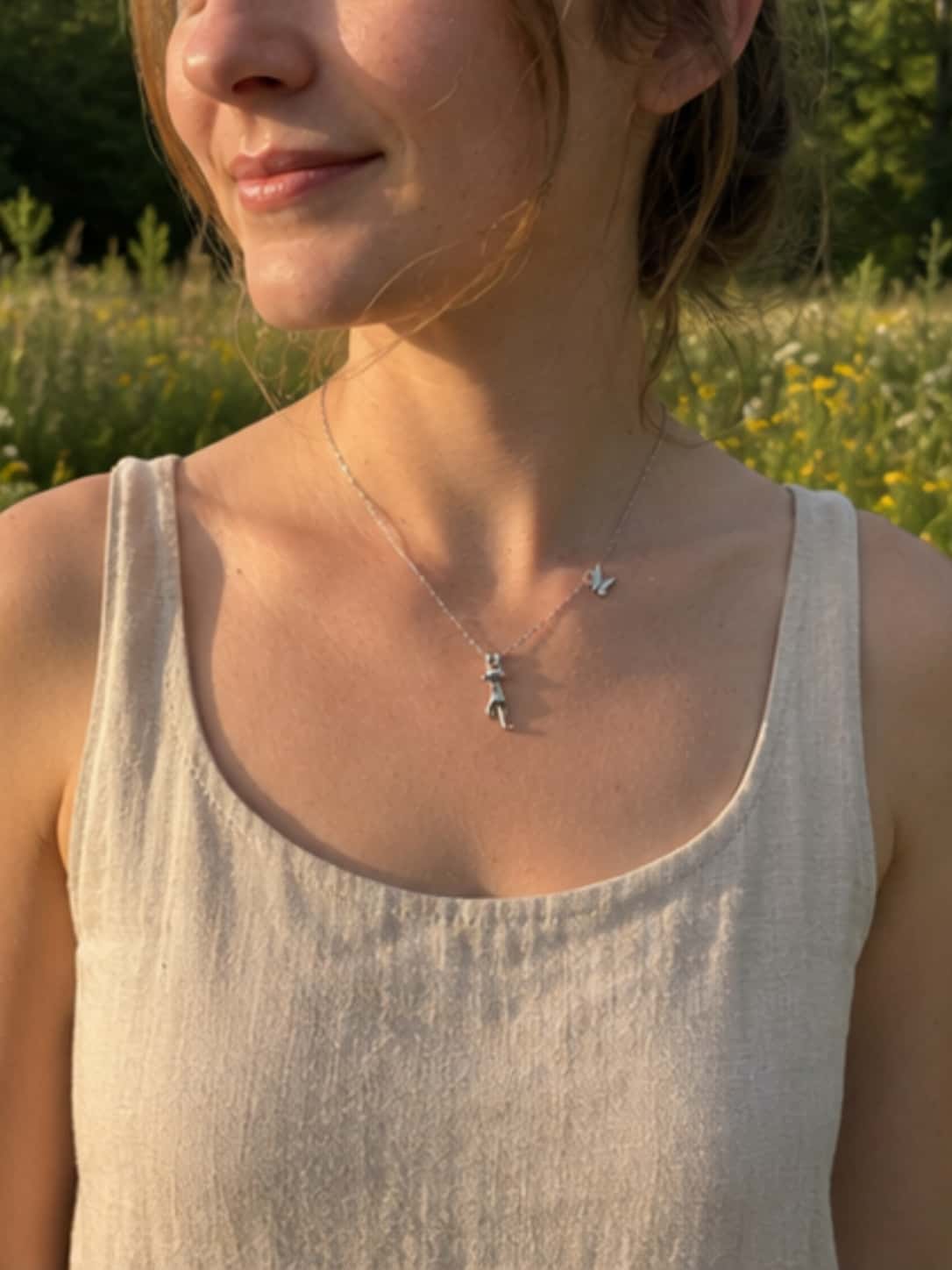 Model wearing a dainty S925 silver cat and butterfly charm necklace paired with a beige linen top, outdoors in a sunlit wildflower field