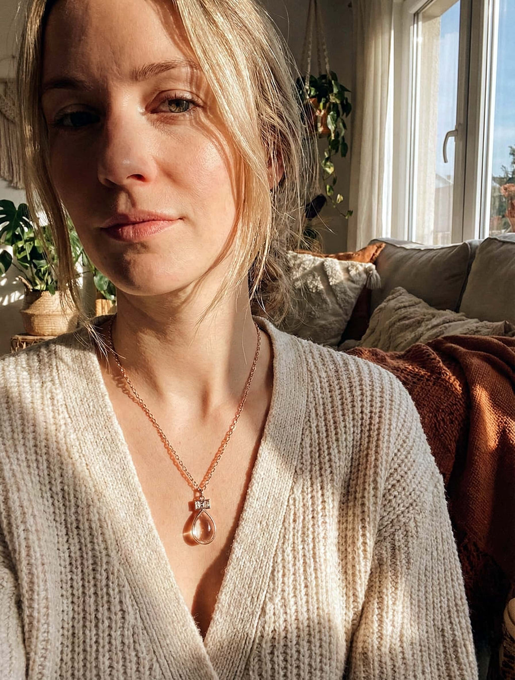 Woman wearing a beige sweater and necklace in a cozy living room.