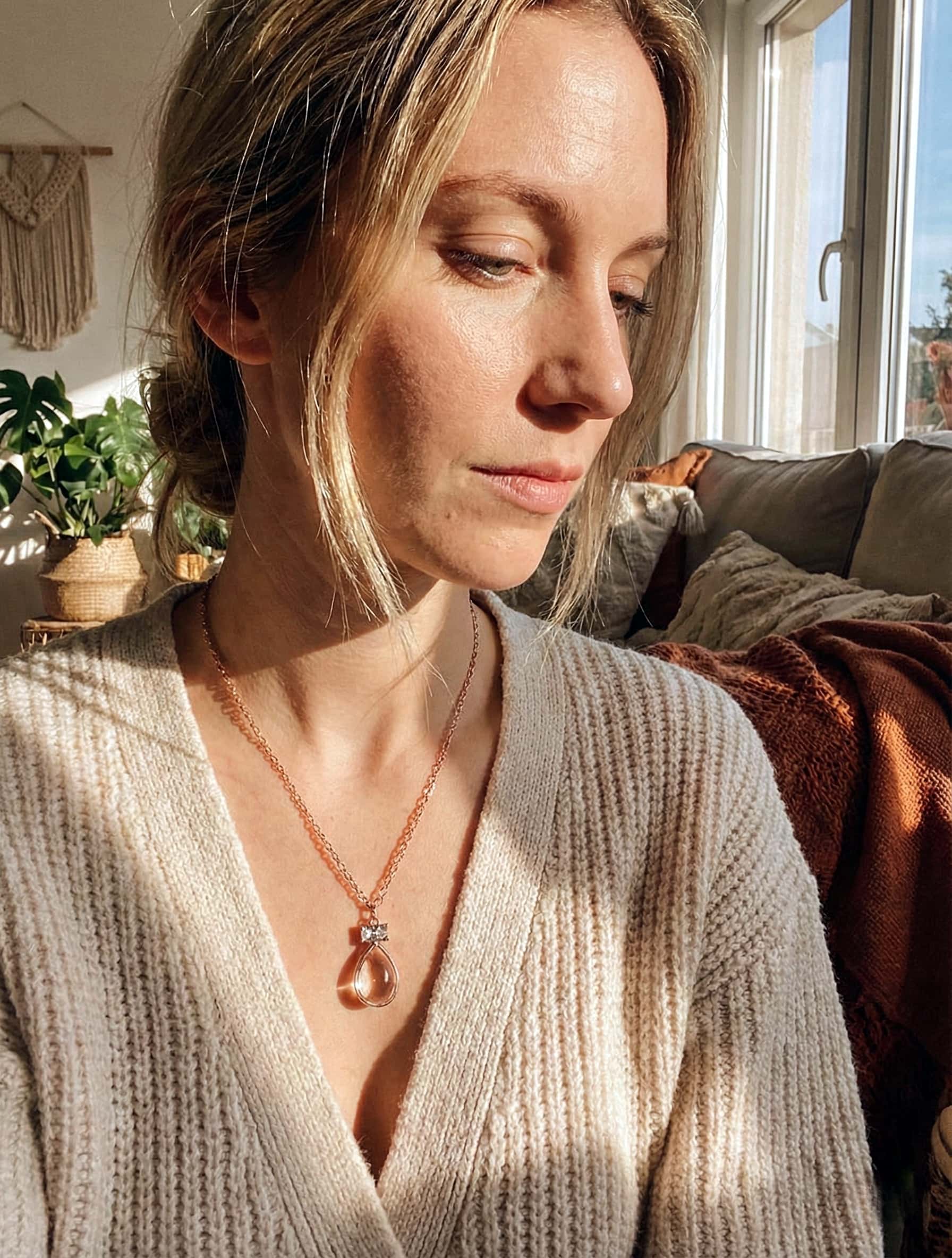 Woman wearing a beige sweater and necklace in a cozy living room.