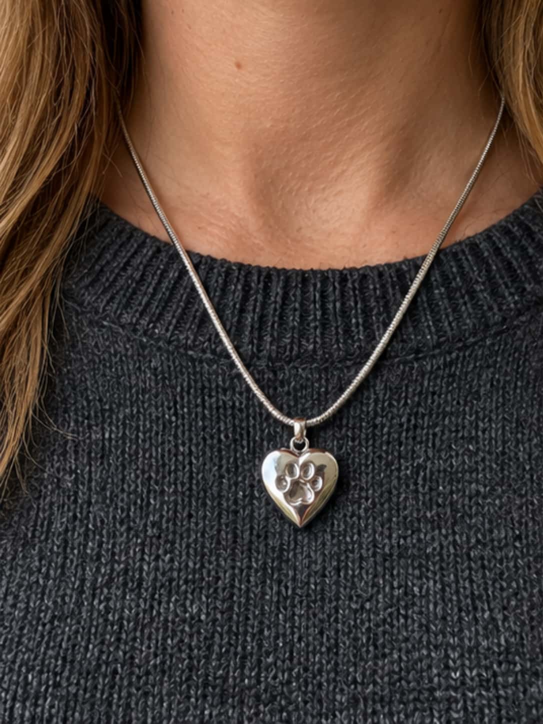 Close-up of a woman wearing a heart-shaped paw print dog ashes necklace over a charcoal knit sweater
