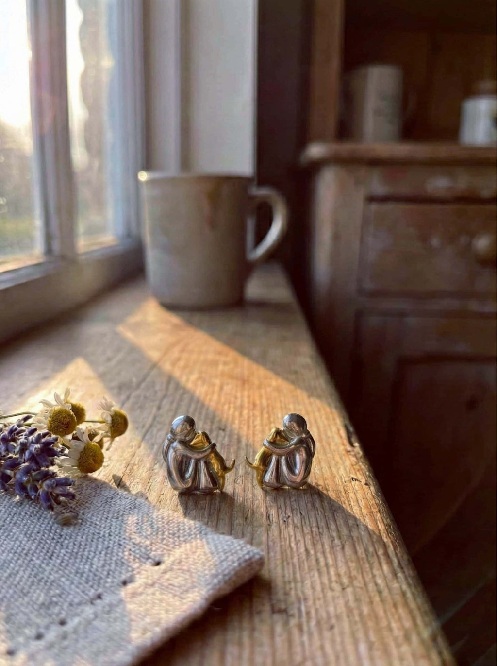 Two small human hug dog earrings on a wooden surface with a mug and lavender in the background.