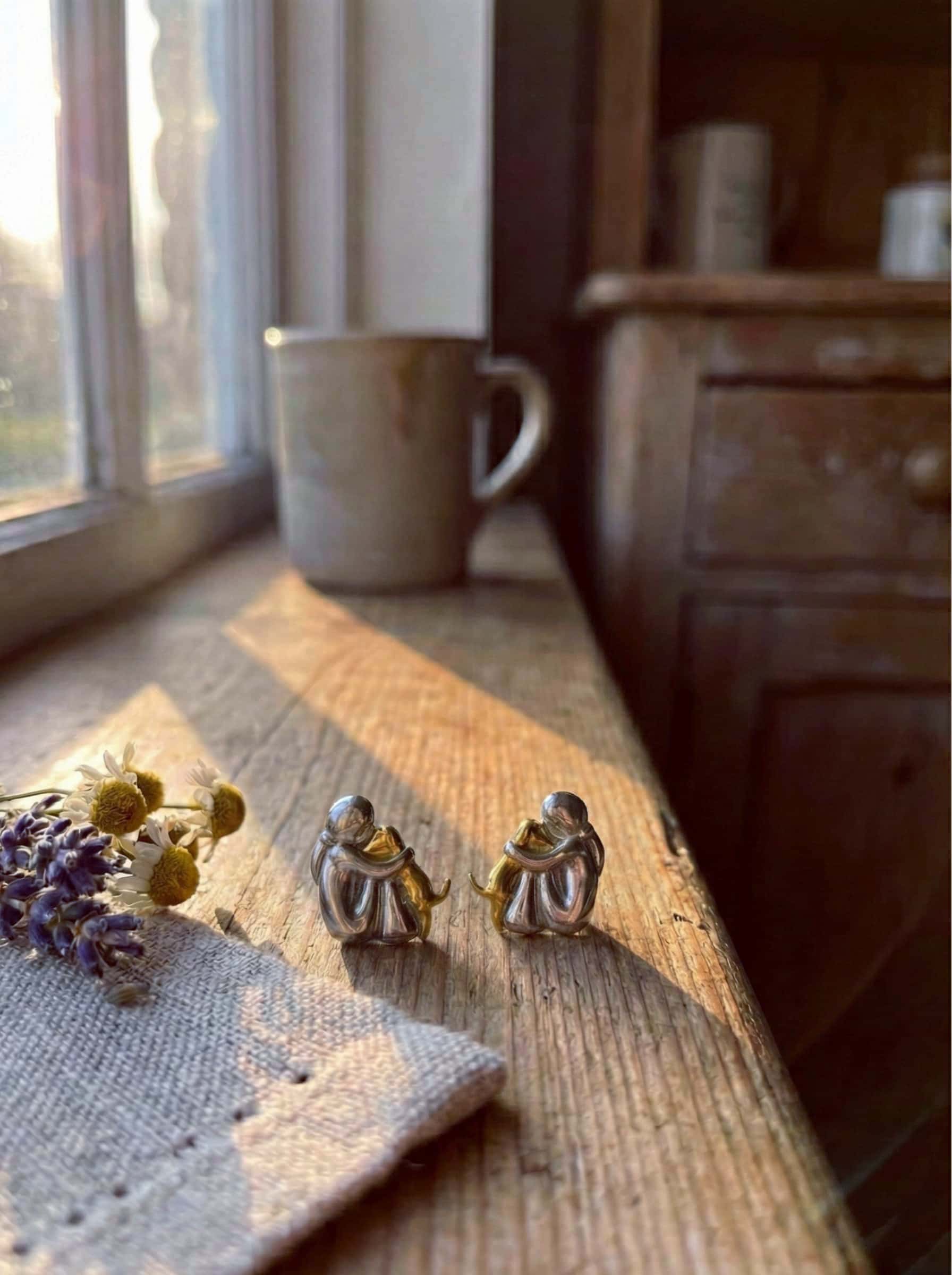 Two small human hug dog earrings on a wooden surface with a mug and lavender in the background.