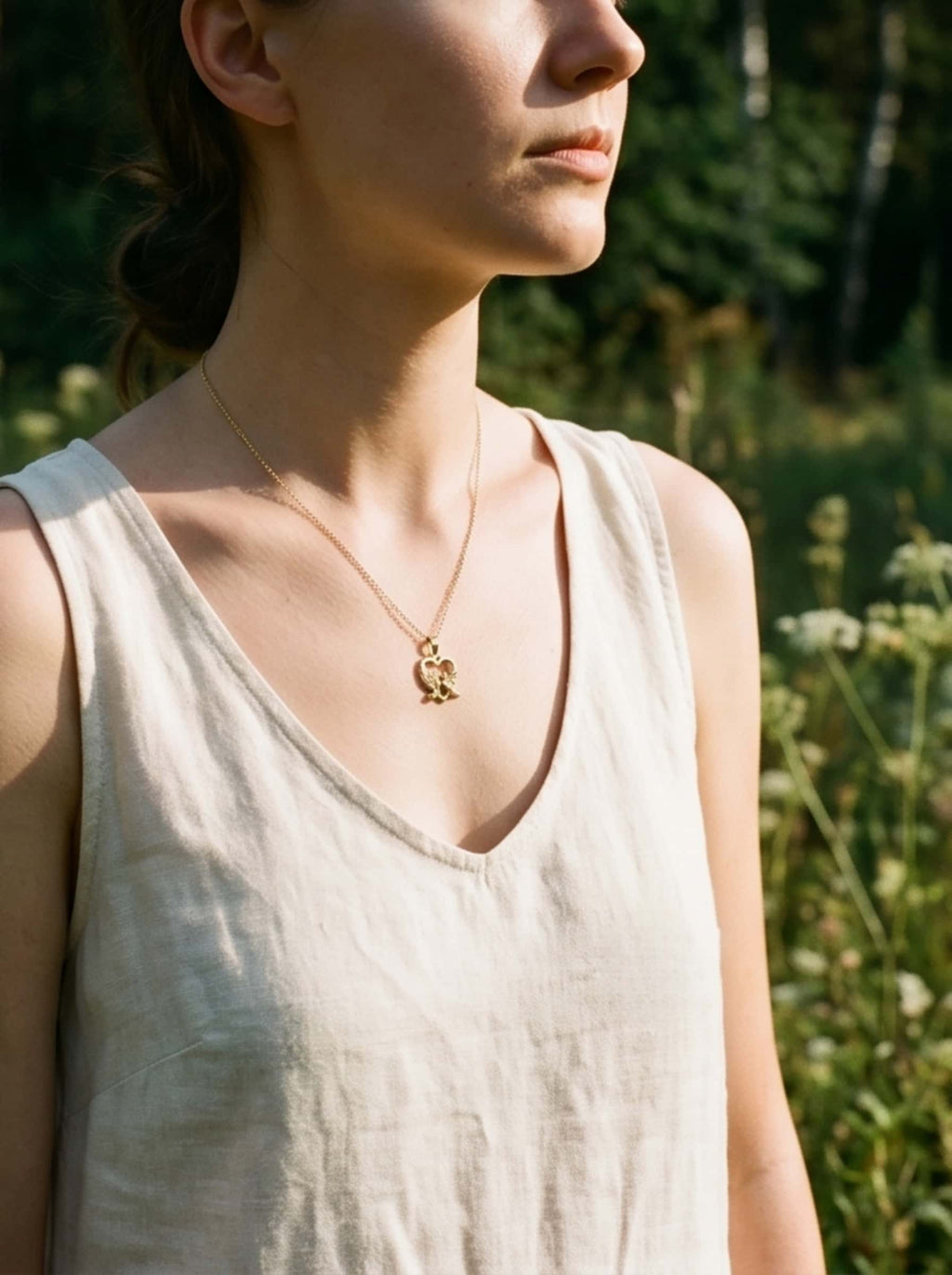 Side profile of a woman outdoors in warm natural sunlight, showcasing the delicate drape and fit of the gold dove pendant necklace against her neckline