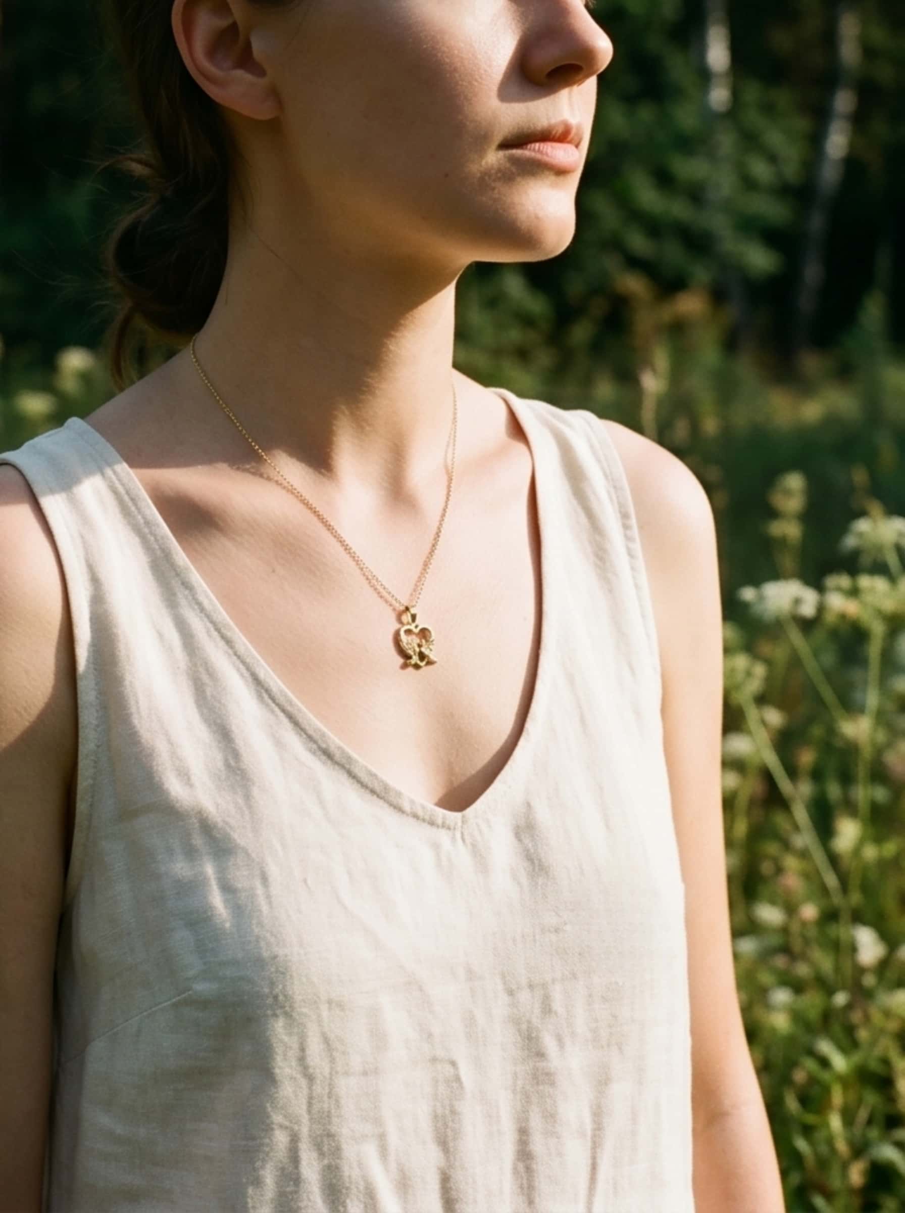 Side profile of a woman outdoors in warm natural sunlight, showcasing the delicate drape and fit of the gold dove pendant necklace against her neckline