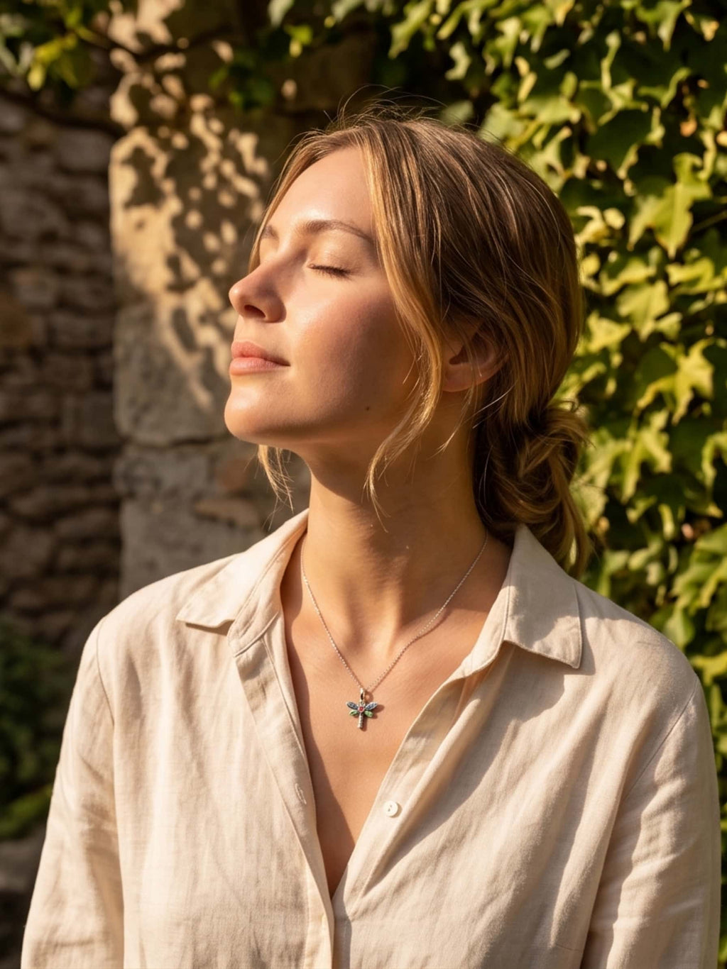 A woman standing in warm sunlight with her eyes closed, wearing a beautiful enamel dragonfly necklace that perfectly complements her casual linen shirt