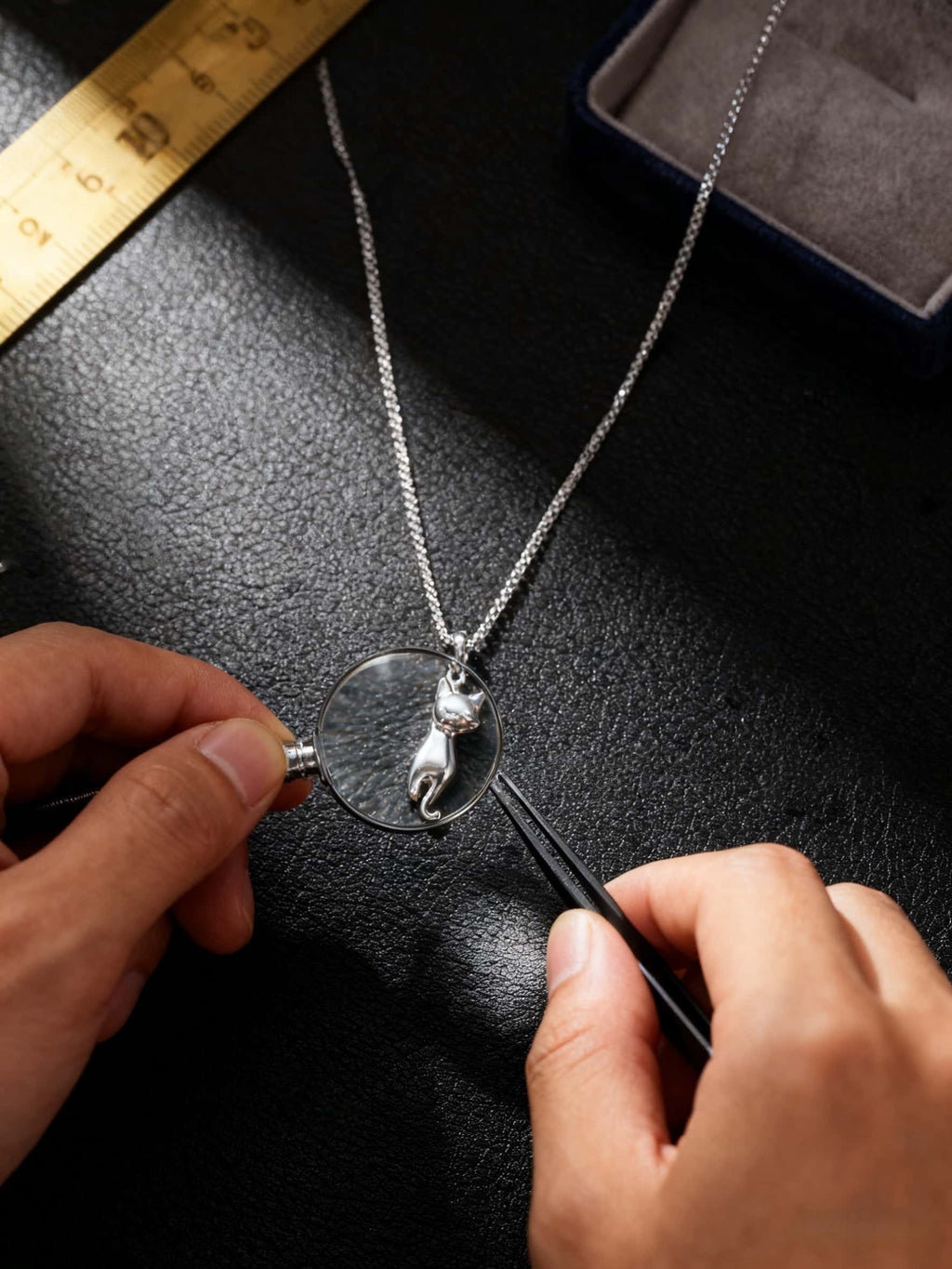 Person examining a silver cat-shaped necklace with a magnifying glass on a dark surface