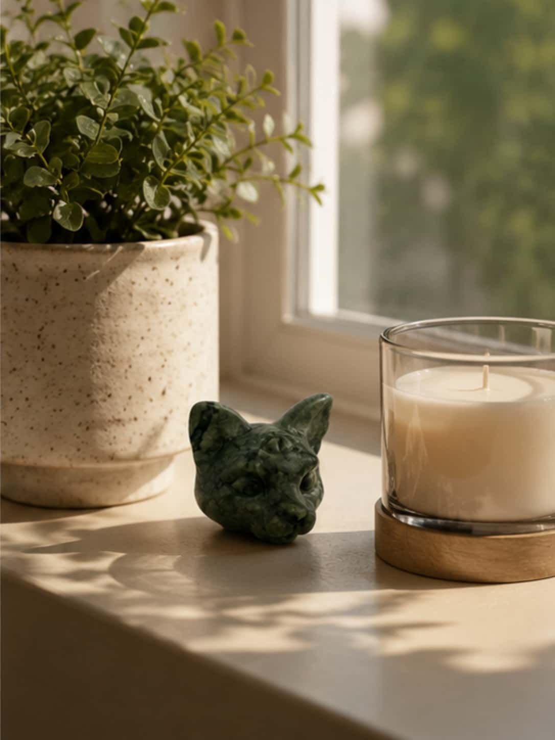 Natural crystal cat figurine sitting on a sunlit windowsill next to a scented glass candle and potted greenery