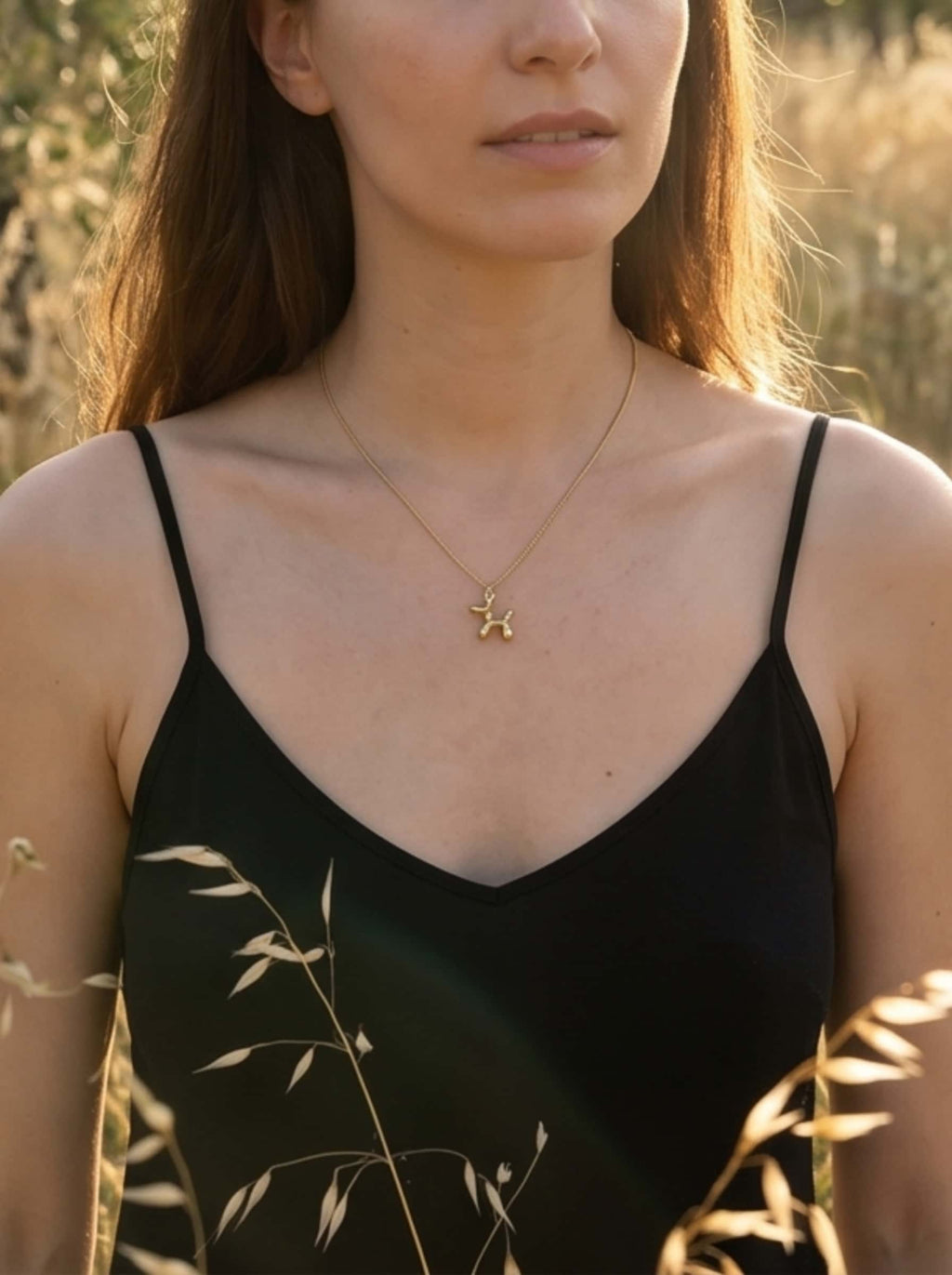 A lifestyle shot of a model wearing the gold balloon dog pendant necklace outdoors in natural, warm golden hour sunlight, styled with a minimalist black top