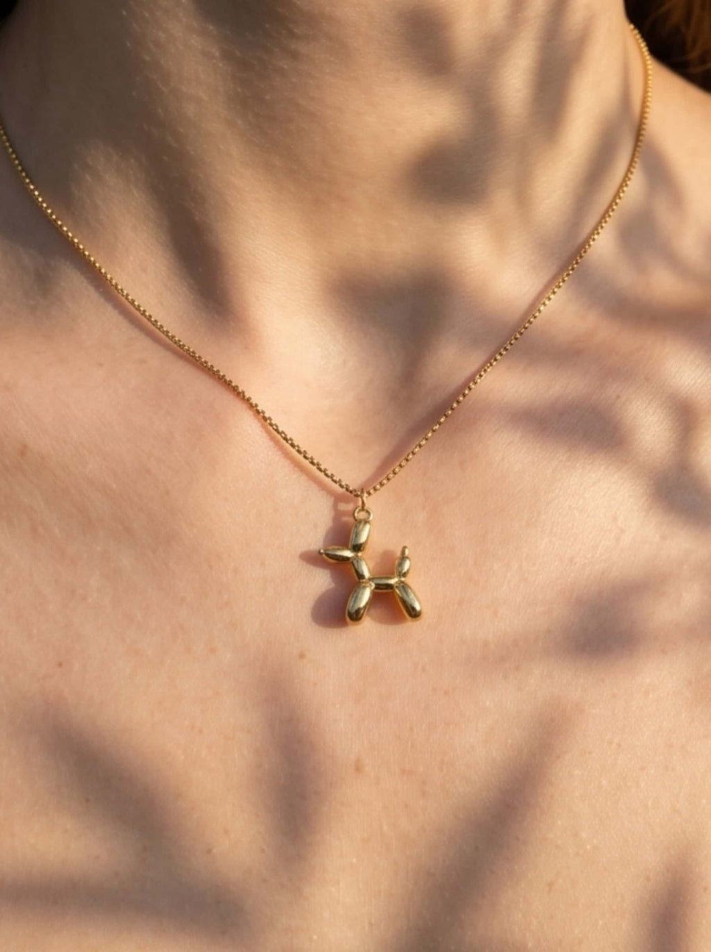 A macro detailed shot of the shiny 18K gold plated balloon dog pendant resting gracefully on skin under artistic, dappled leaf shadows