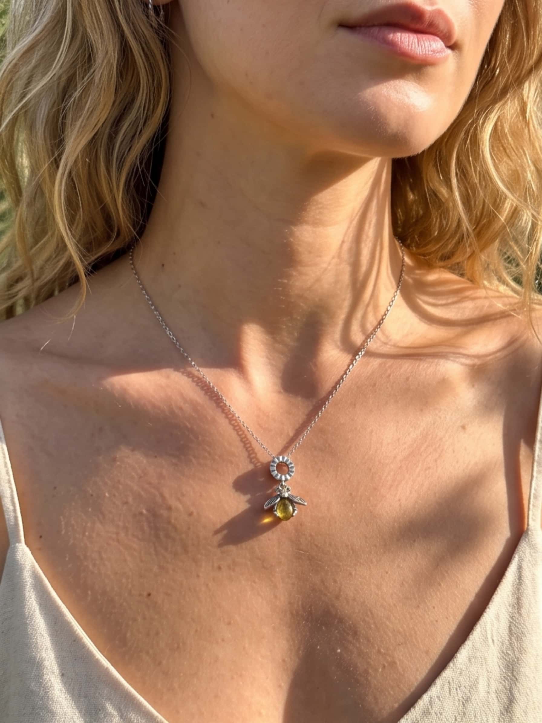 Close-up of a woman's décolletage wearing a gold bee pendant necklace style featuring a glowing yellow crystal and silver wings, illuminated by warm sunlight