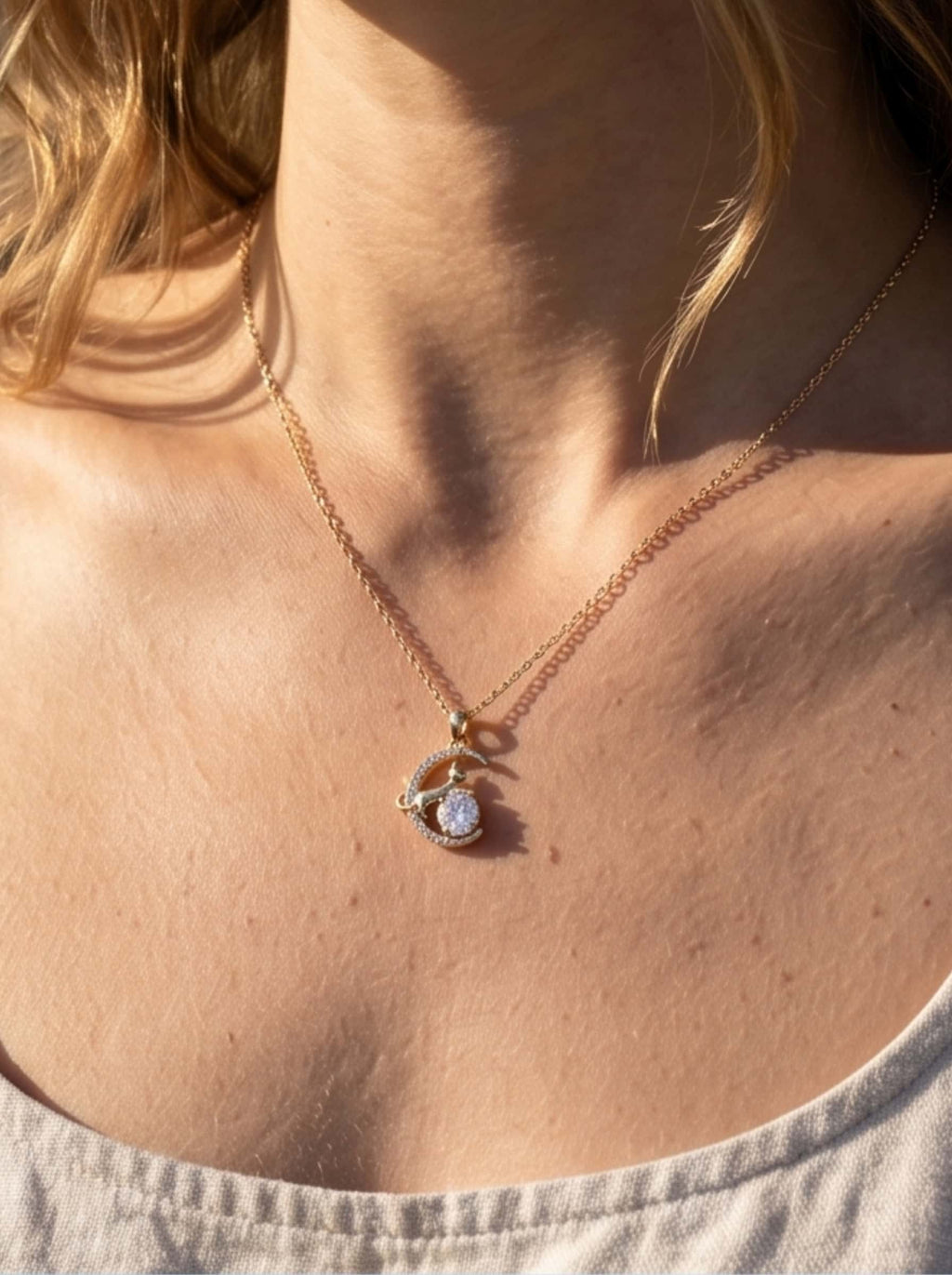 Close-up detail of a delicate gold celestial cat pendant necklace worn by a woman, illuminated by warm, natural sunlight showcasing the radiant 18K gold plating
