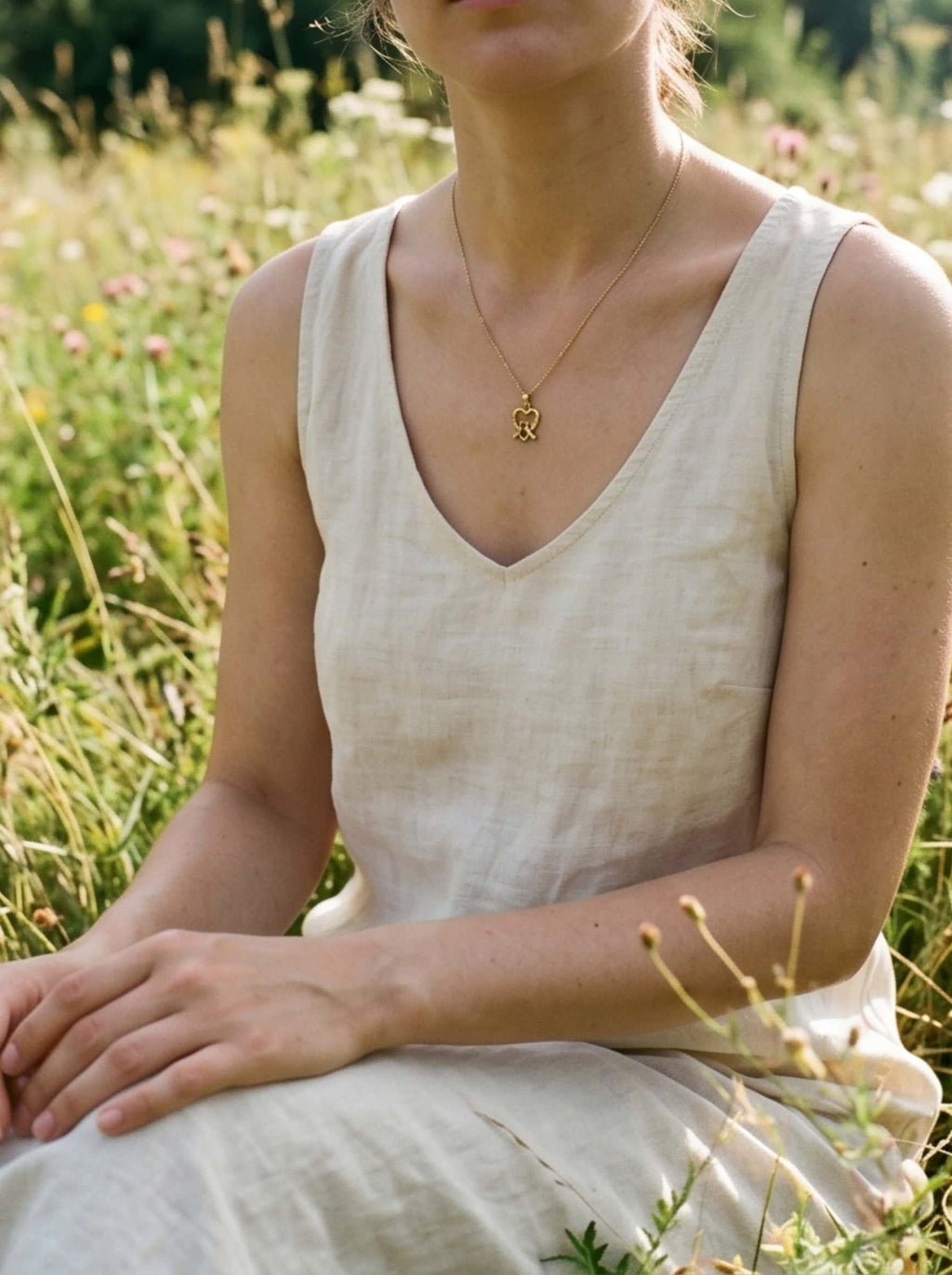 Mid-shot of a female model sitting in a sunny wildflower field, wearing a cream linen sleeveless dress paired with an elegant gold dove necklace