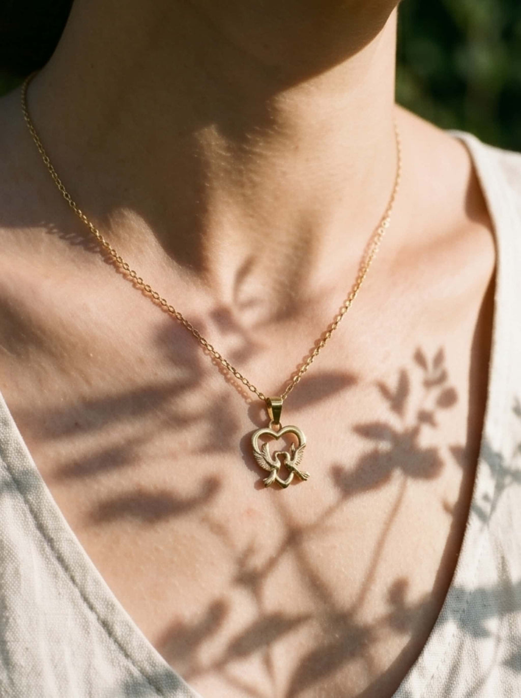 Aesthetic close-up macro shot of the gold dove pendant necklace on a model’s chest, with artistic, soft botanical shadows of leaves dancing across her skin and linen dress