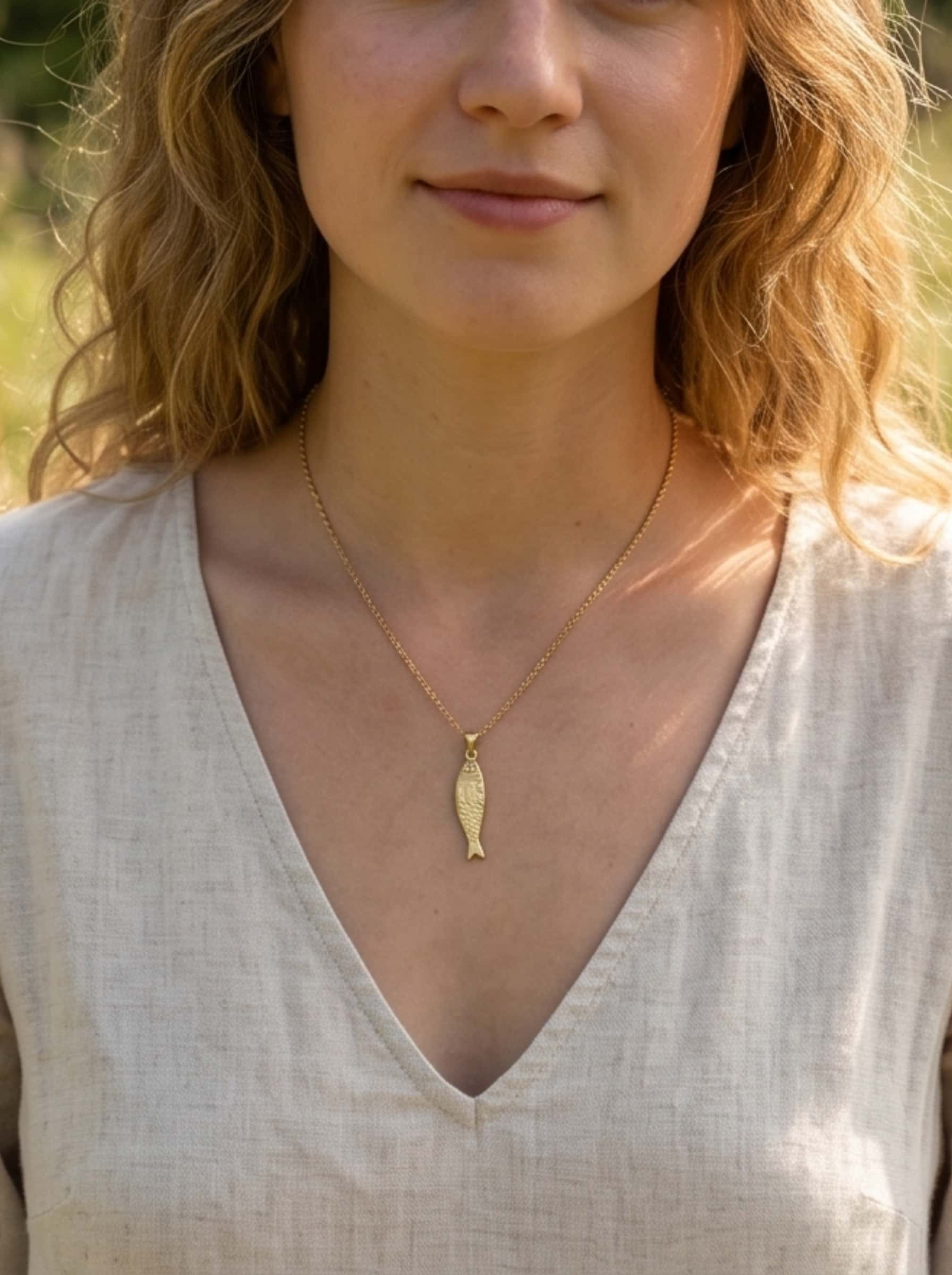 A blonde woman in a beige organic linen V-neck top wearing the delicate gold fish necklace, captured in warm, natural outdoor sunlight