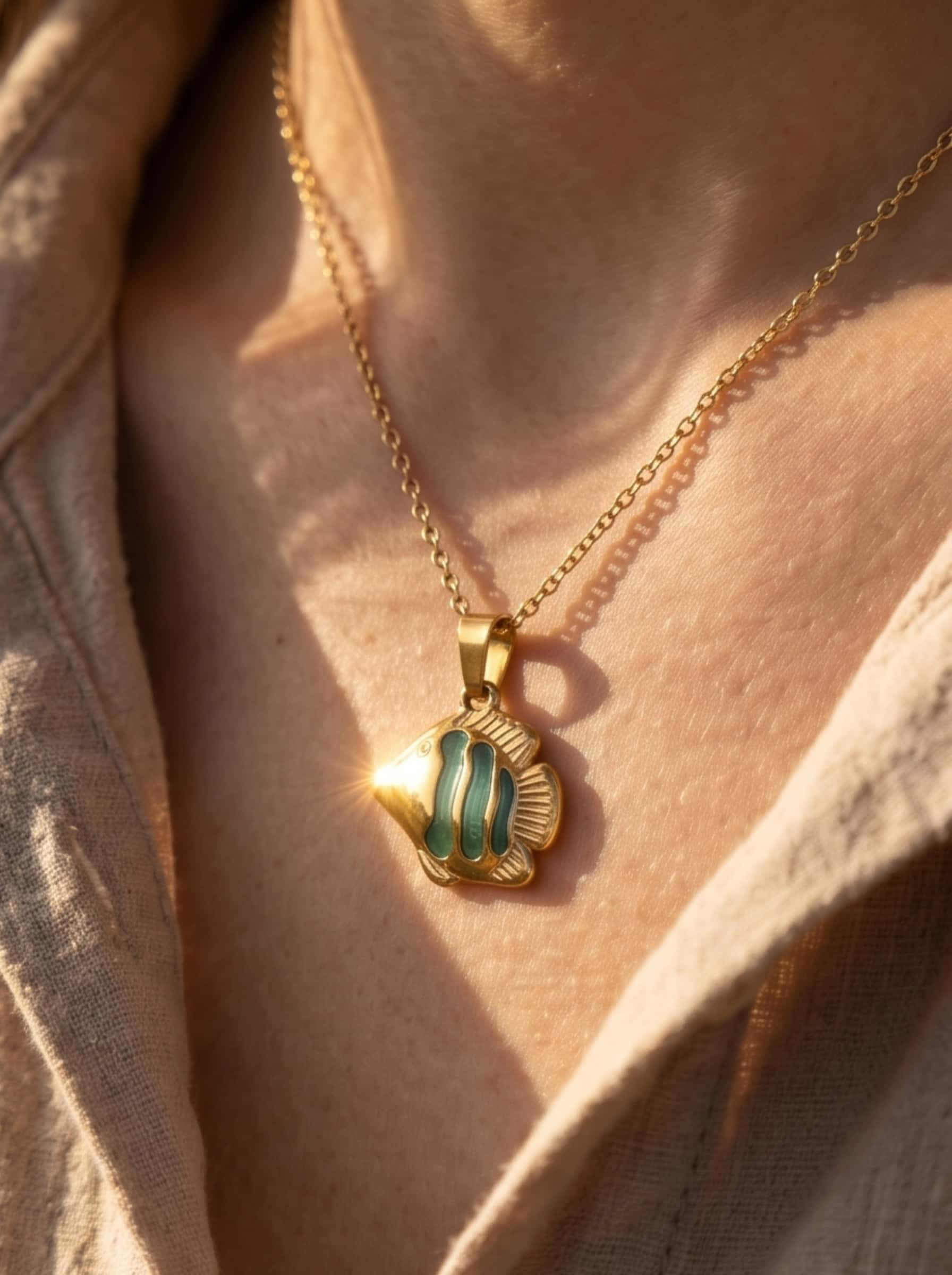 A sun-drenched close-up of the enamel angelfish pendant necklace glistening on a model's neck, capturing a natural sun flare and the texture of an organic linen shirt