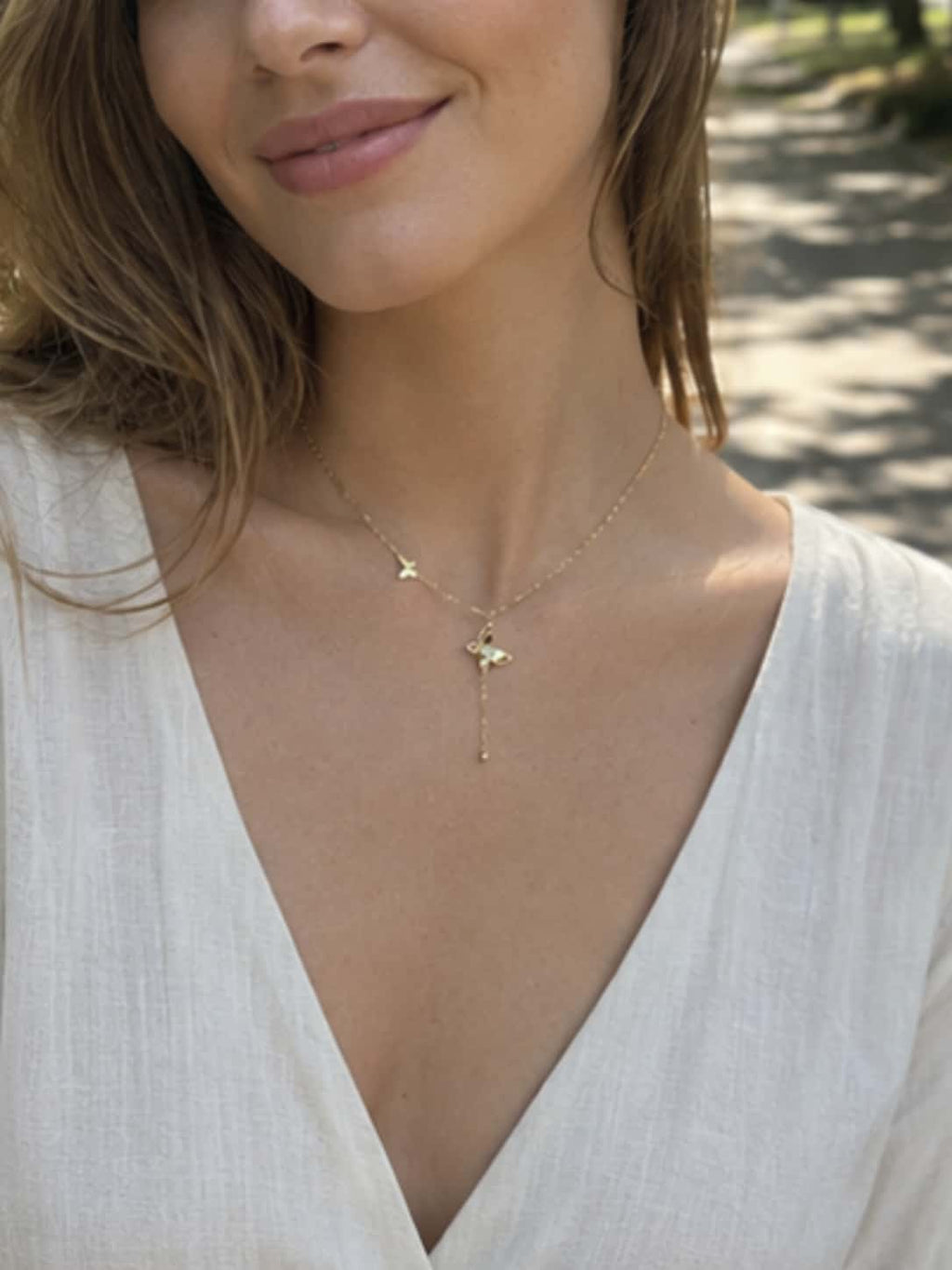 A smiling female model wearing a white linen V-neck dress and the dainty gold necklace butterfly lifestyle shot outdoors in soft morning light