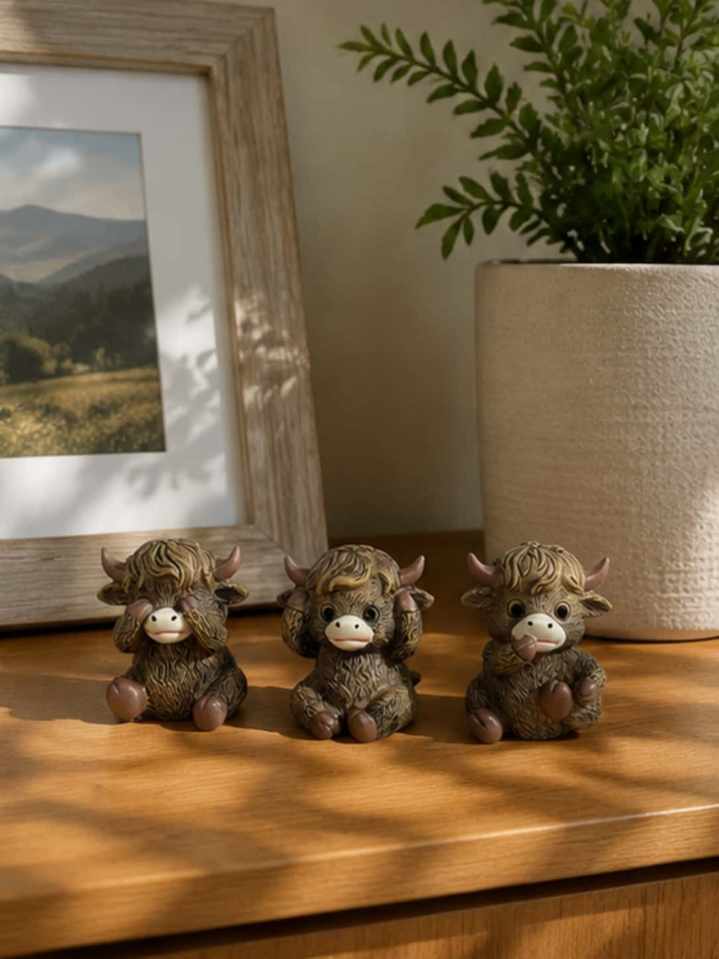 Three-piece highland cow decor set styled on a natural wooden tabletop next to a potted plant and framed art