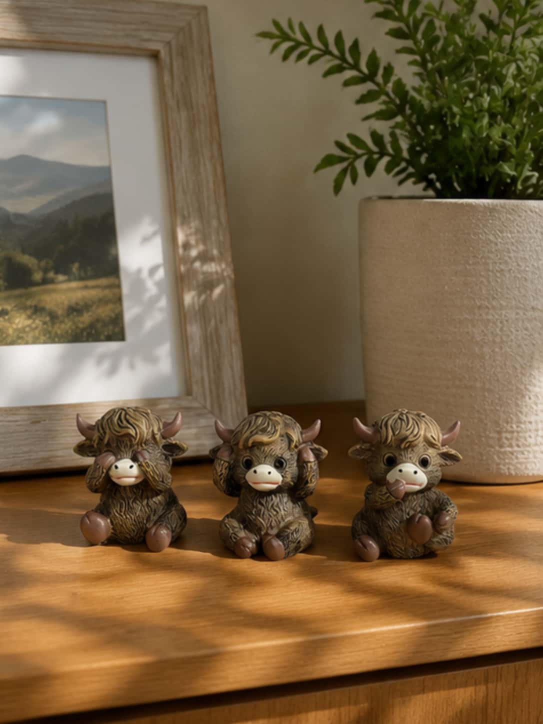 Three-piece highland cow decor set styled on a natural wooden tabletop next to a potted plant and framed art