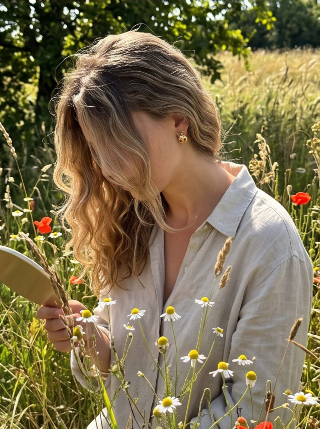 Lifestyle shot of a model wearing a relaxed linen shirt and 18K gold ladybug earrings holding a handheld mirror in a field of blooming daisies