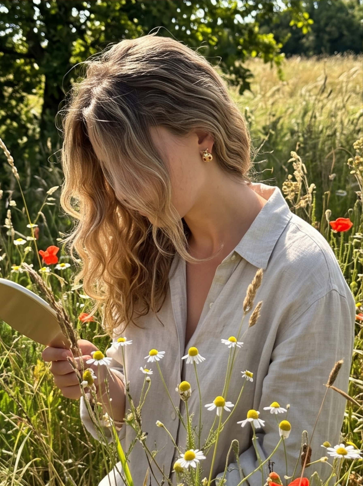 Lifestyle shot of a model wearing a relaxed linen shirt and 18K gold ladybug earrings holding a handheld mirror in a field of blooming daisies