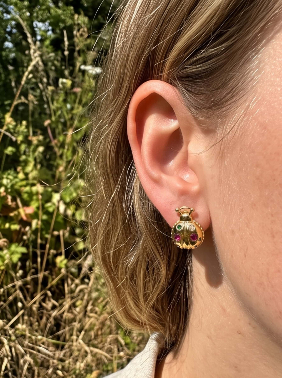 Close-up of a woman wearing the 18K gold plated ladybug earrings handset with ruby-red and emerald-green cubic zirconia in outdoor natural light