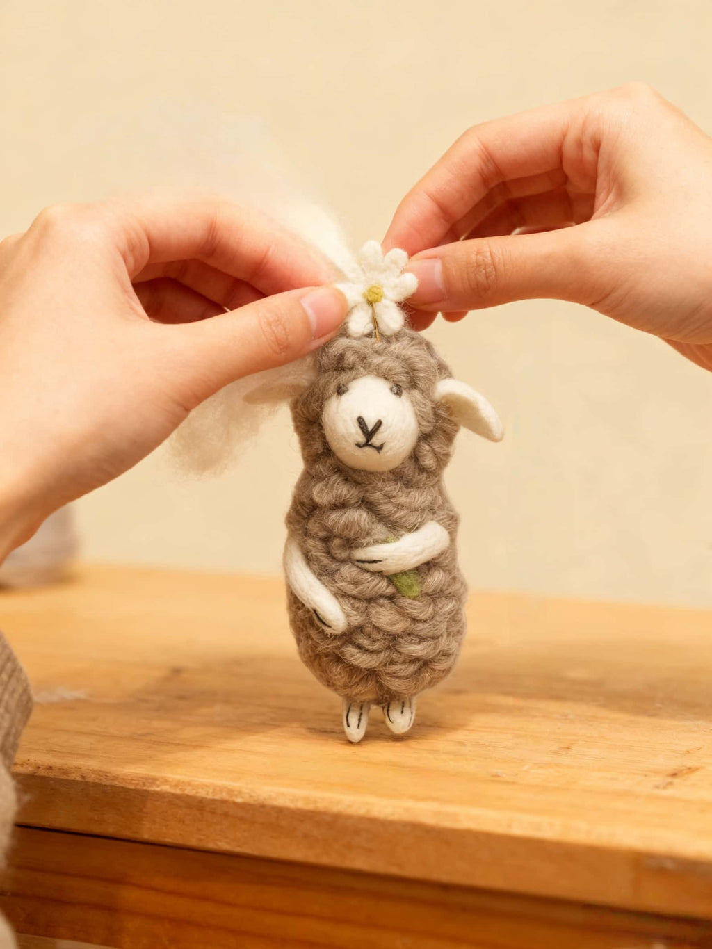 Small felted sheep ornament with a flower held by two hands on a wooden surface.