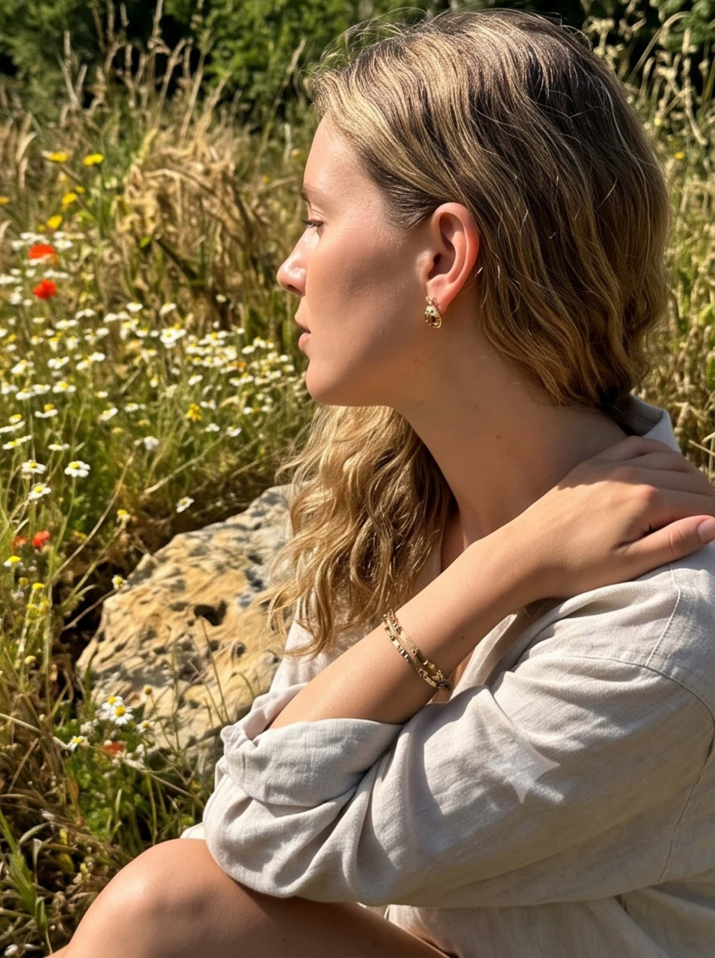 Side profile of a woman wearing gold ladybug earrings and matching bracelets sitting gracefully in a sunny wildflower meadow