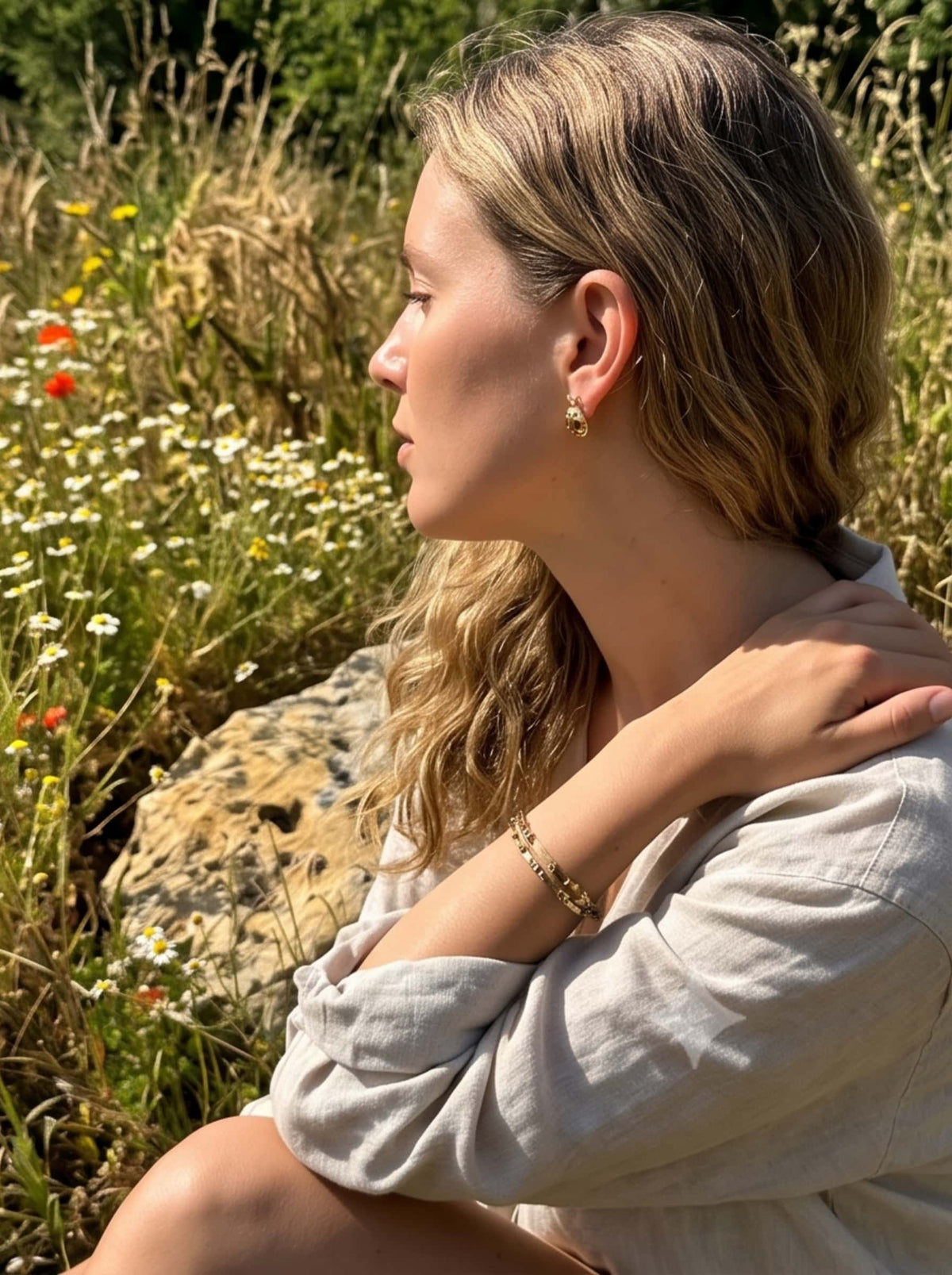 Side profile of a woman wearing gold ladybug earrings and matching bracelets sitting gracefully in a sunny wildflower meadow