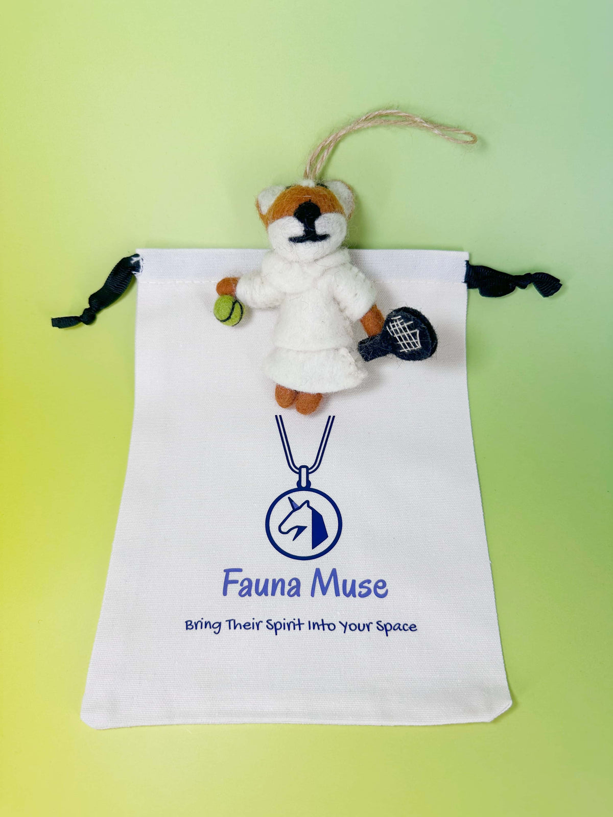 Handmade needle felted wool fox ornament wearing a white tennis outfit holding a racket and ball with Fauna Muse gift pouch