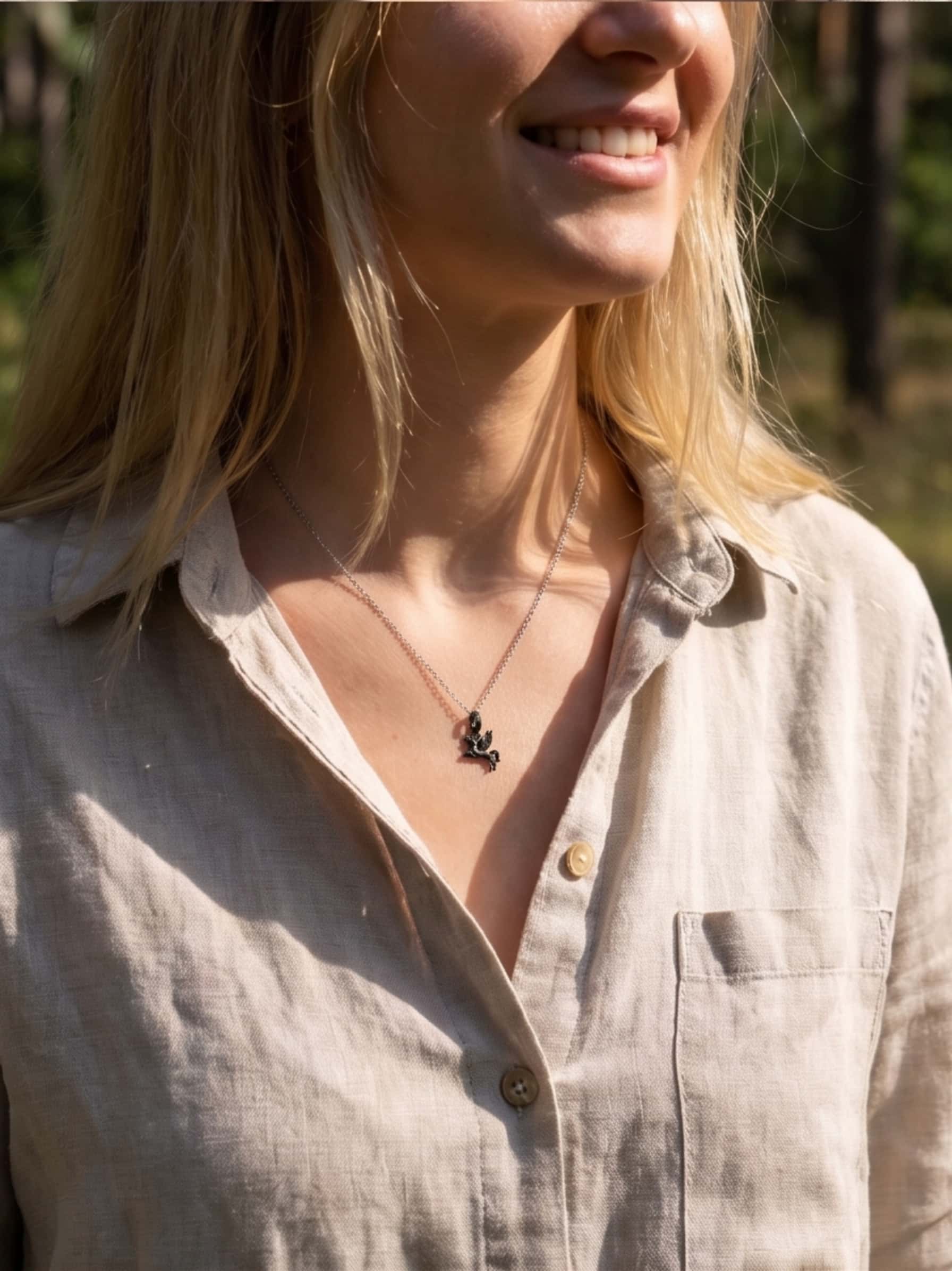 A smiling woman outdoors wearing a beige button-down shirt paired perfectly with the black rhodium winged unicorn necklace, demonstrating casual everyday styling