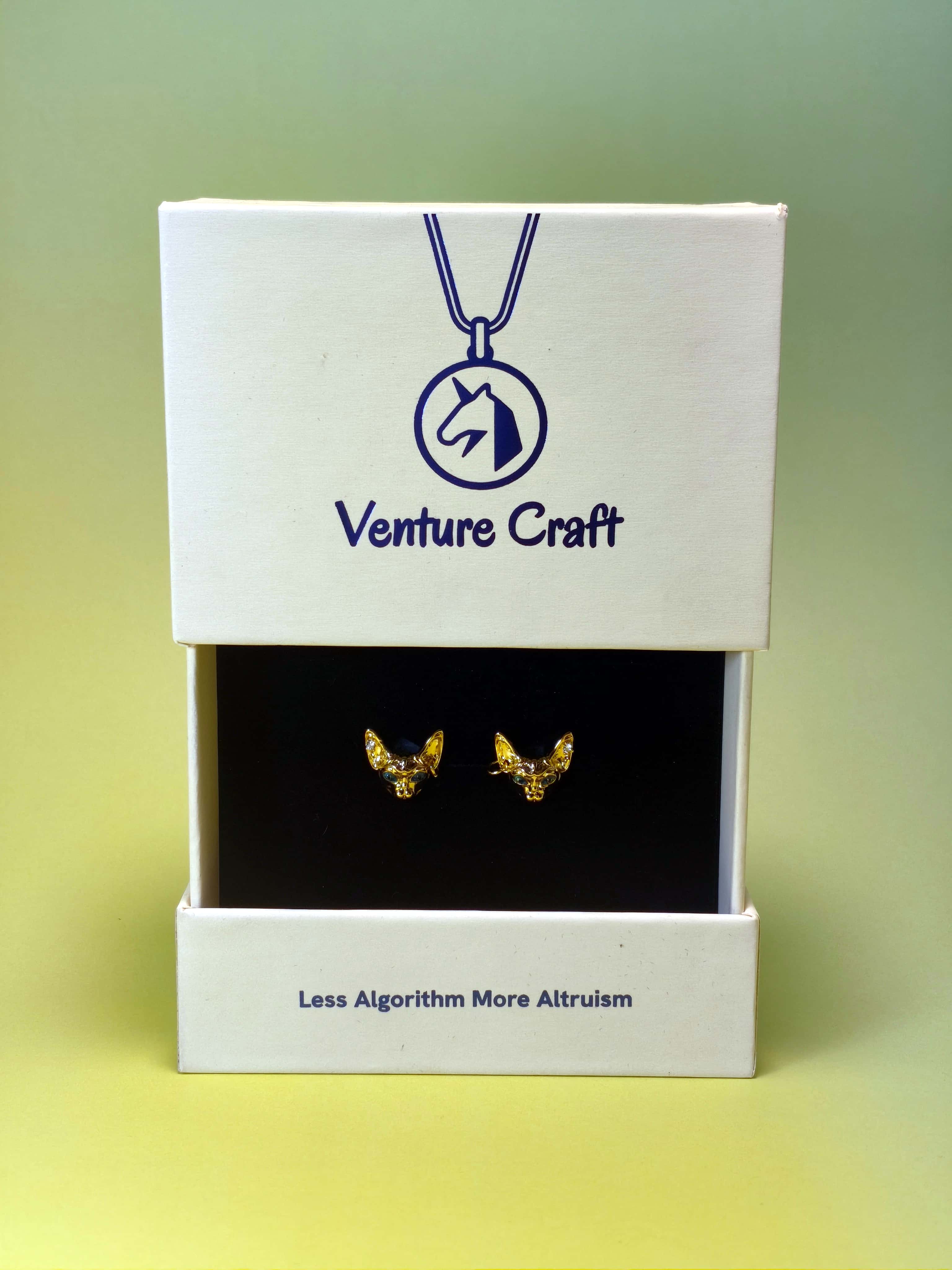 Gold sphinx-cat earrings in a Venture Craft gift box on a gradient background