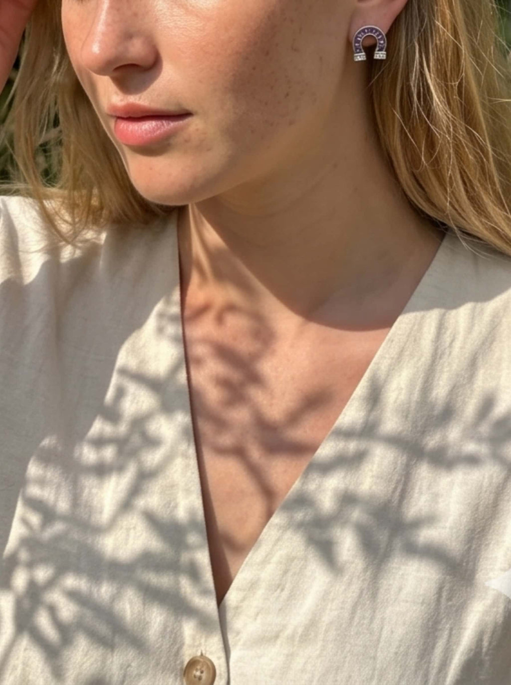 A close-up view of a natural-lit blonde model wearing a sparkling diamond horseshoe earrings featuring prominent baguette-cut purple stones and micro-pave accents, paired with a beige linen shirt