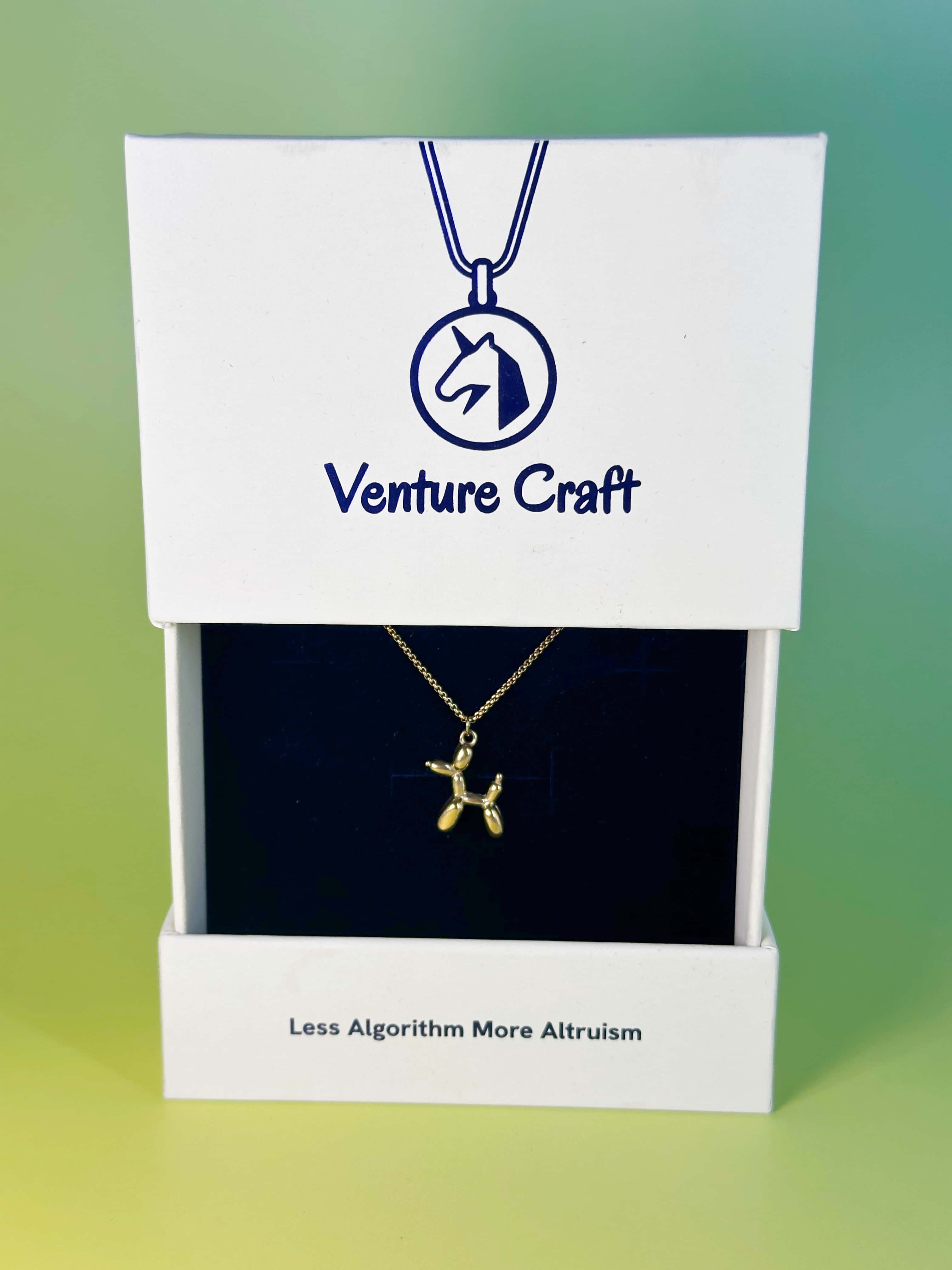 The gold balloon dog pendant necklace beautifully displayed inside its premium white Venture Craft gift box, featuring the brand slogan 'Less Algorithm More Altruism'