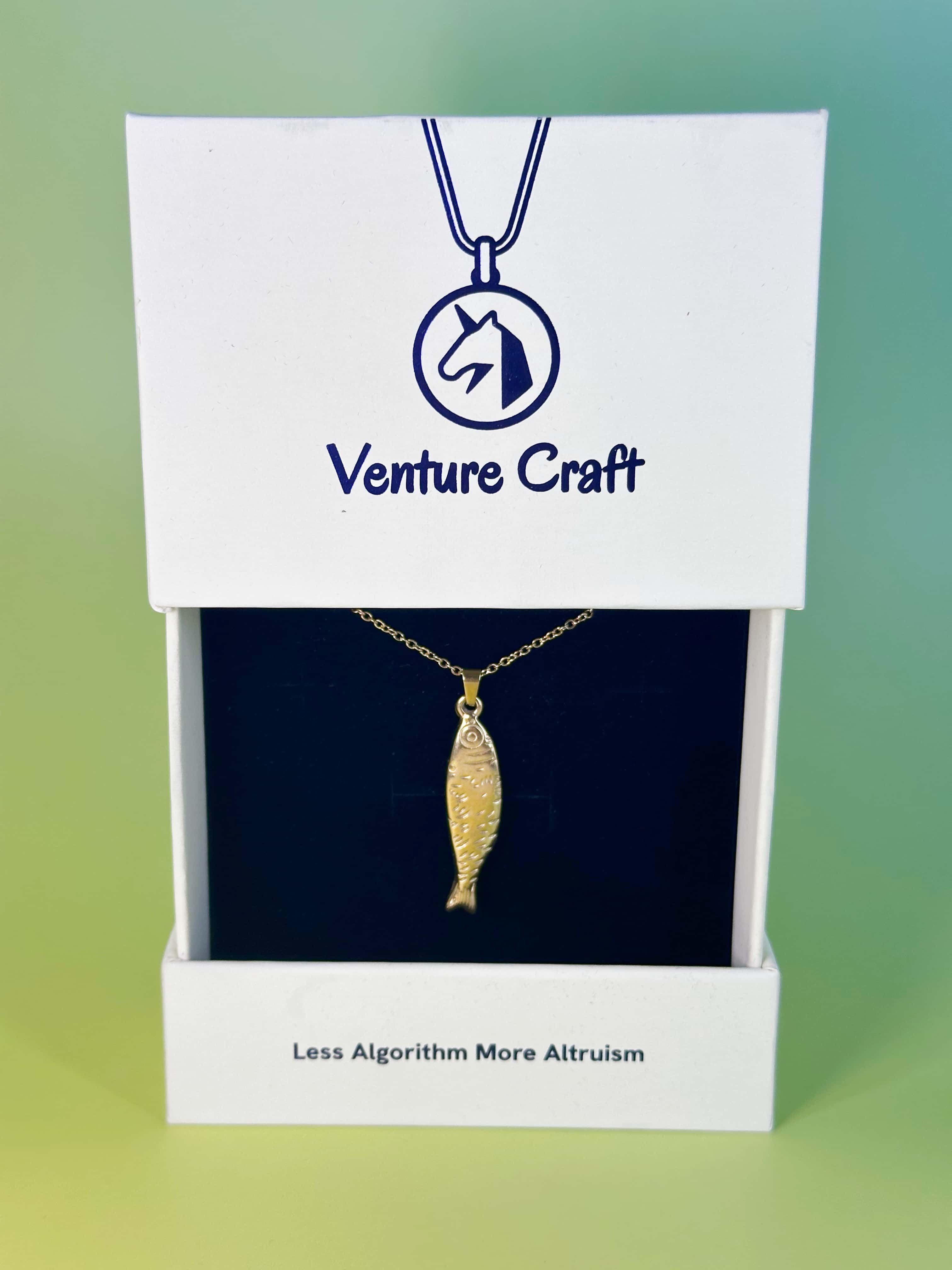 The elegant 18K gold fish pendant necklace displayed inside its sustainable white Venture Craft gift box with the brand motto Less Algorithm More Altruism
