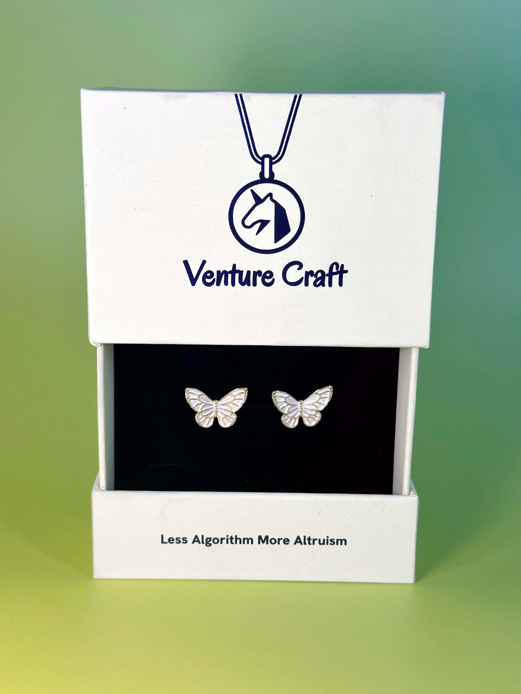 Premium sustainable white jewelry box opening to reveal gold butterfly earrings, featuring the Venture Craft logo and Less Algorithm More Altruism brand slogan