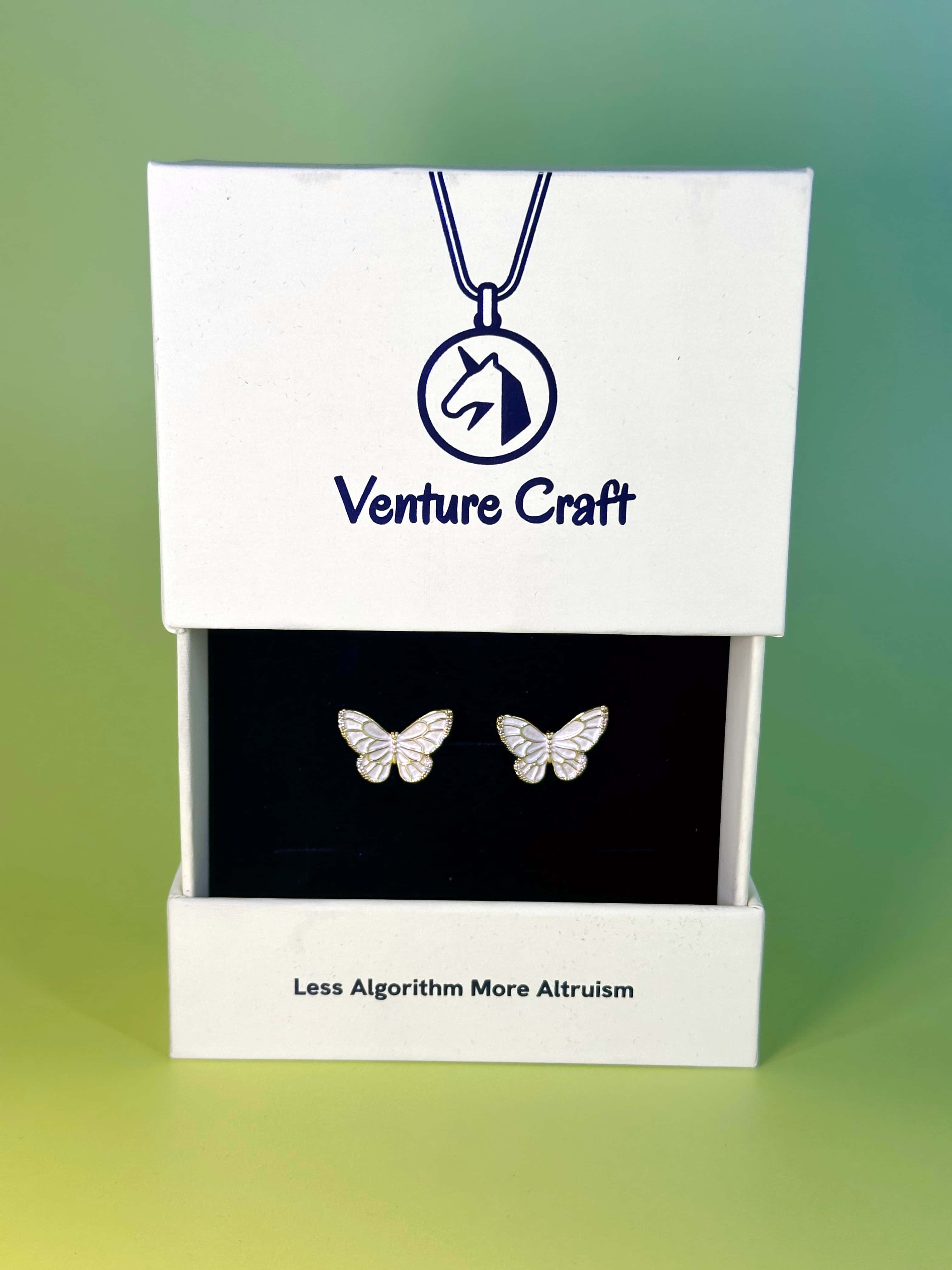 Premium sustainable white jewelry box opening to reveal gold butterfly earrings, featuring the Venture Craft logo and Less Algorithm More Altruism brand slogan