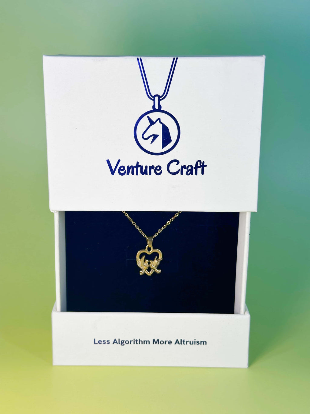 Premium white slide-out packaging box from Venture Craft displaying the gold dove necklace on a dark navy velvet insert, featuring a blue unicorn logo and the brand slogan 'Less Algorithm More Altruism'