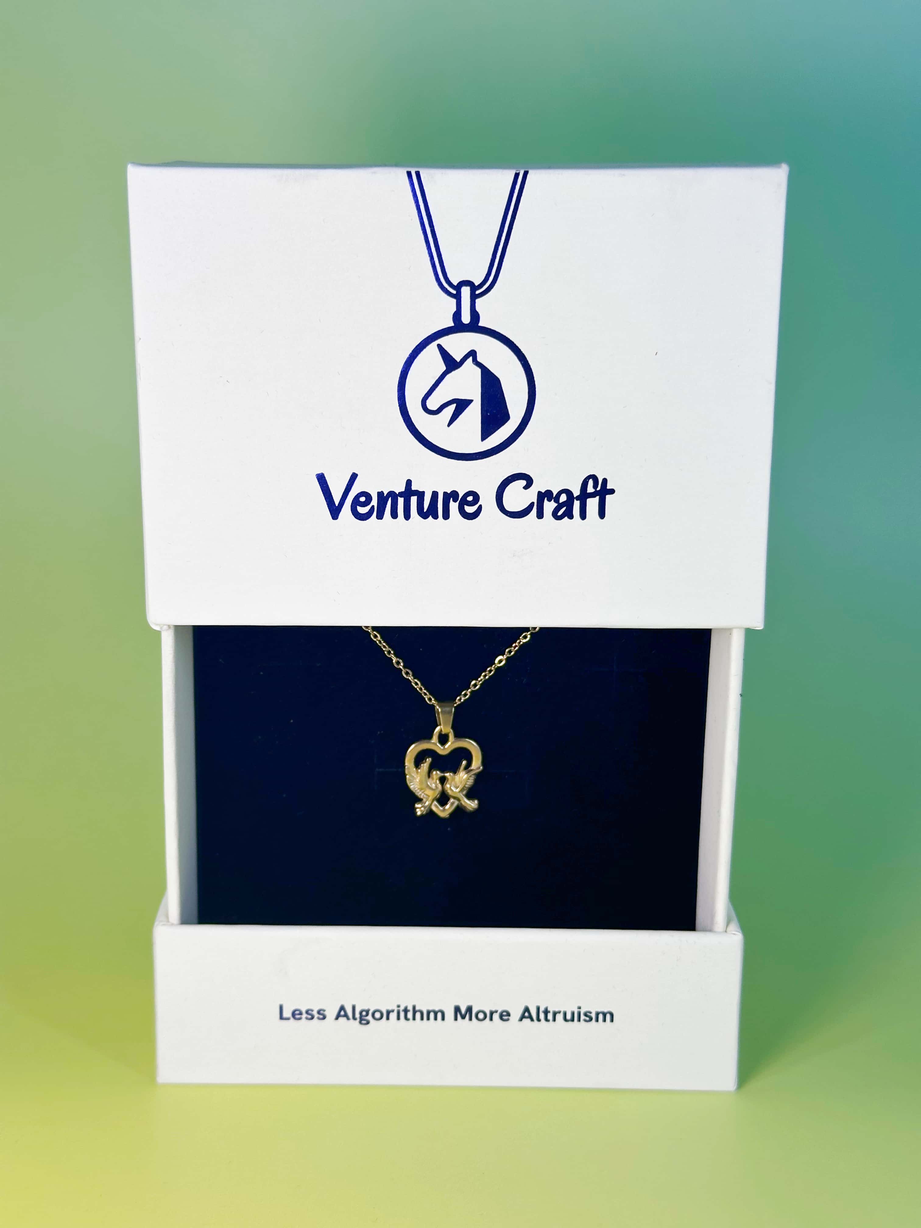 Premium white slide-out packaging box from Venture Craft displaying the gold dove necklace on a dark navy velvet insert, featuring a blue unicorn logo and the brand slogan 'Less Algorithm More Altruism'