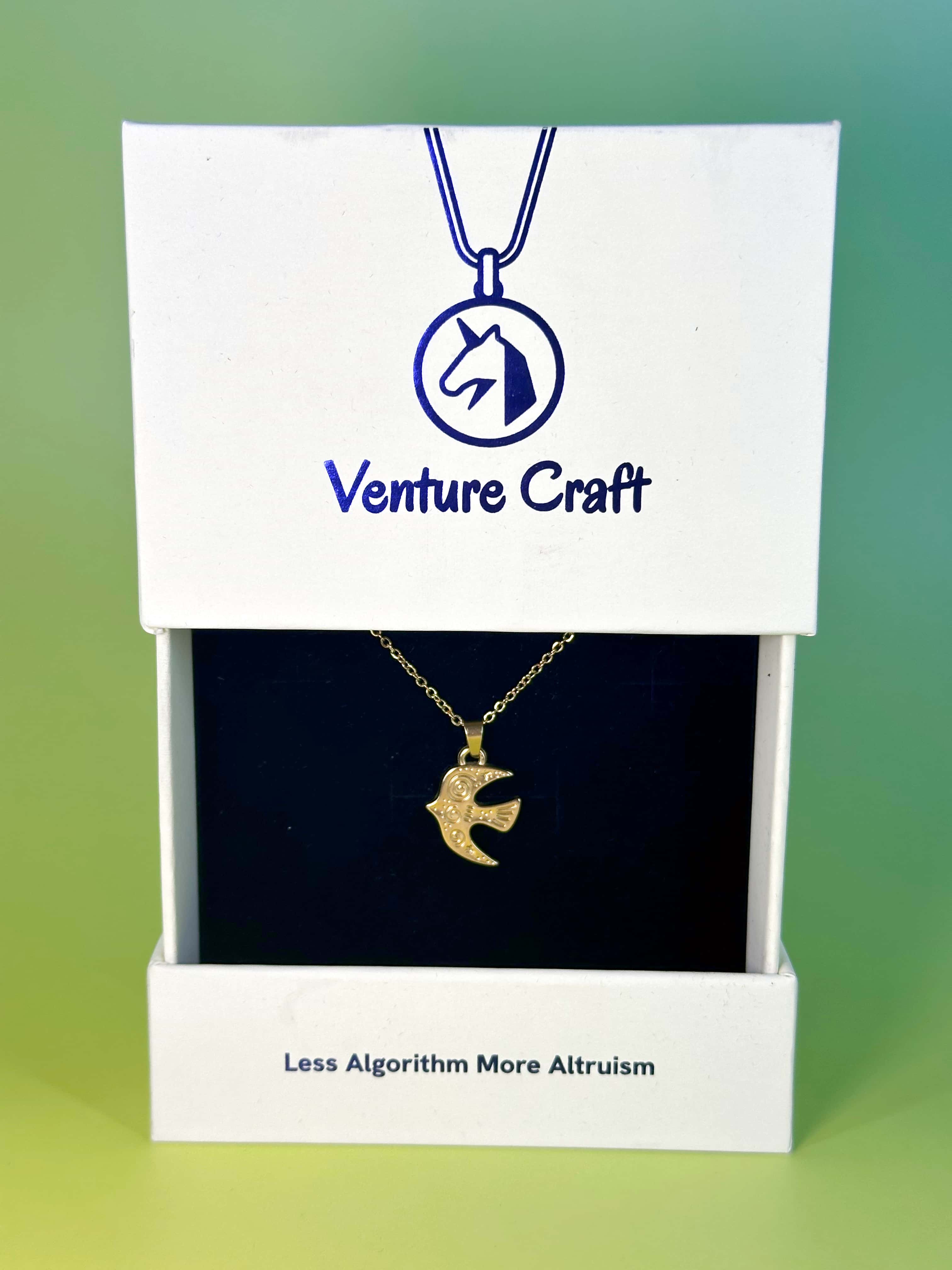 The elegant gold bird necklace displayed inside its white Venture Craft gift box featuring the brand tagline Less Algorithm More Altruism