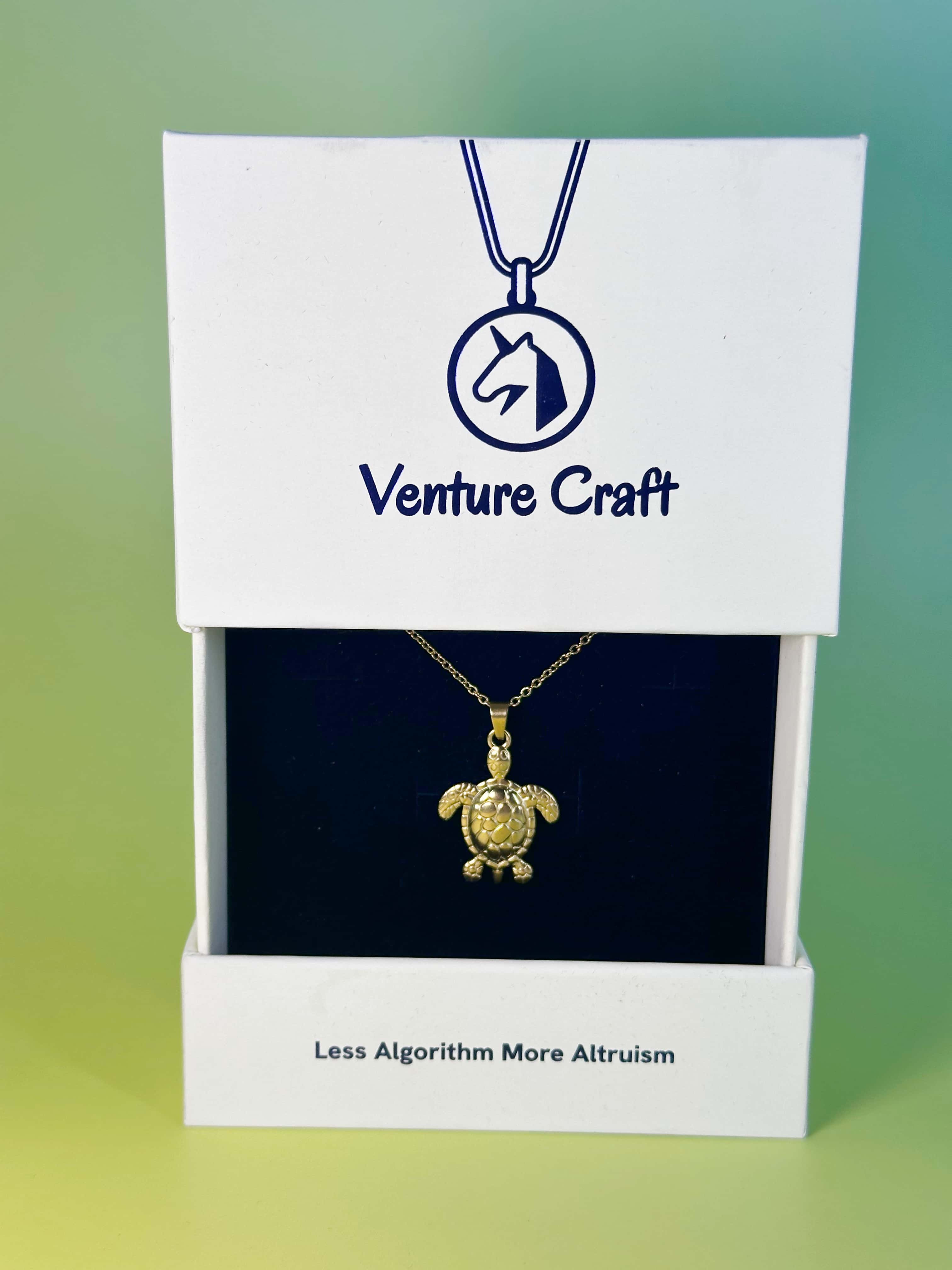 The gold turtle necklace elegantly displayed inside a white Venture Craft branded gift box featuring the slogan Less Algorithm More Altruism