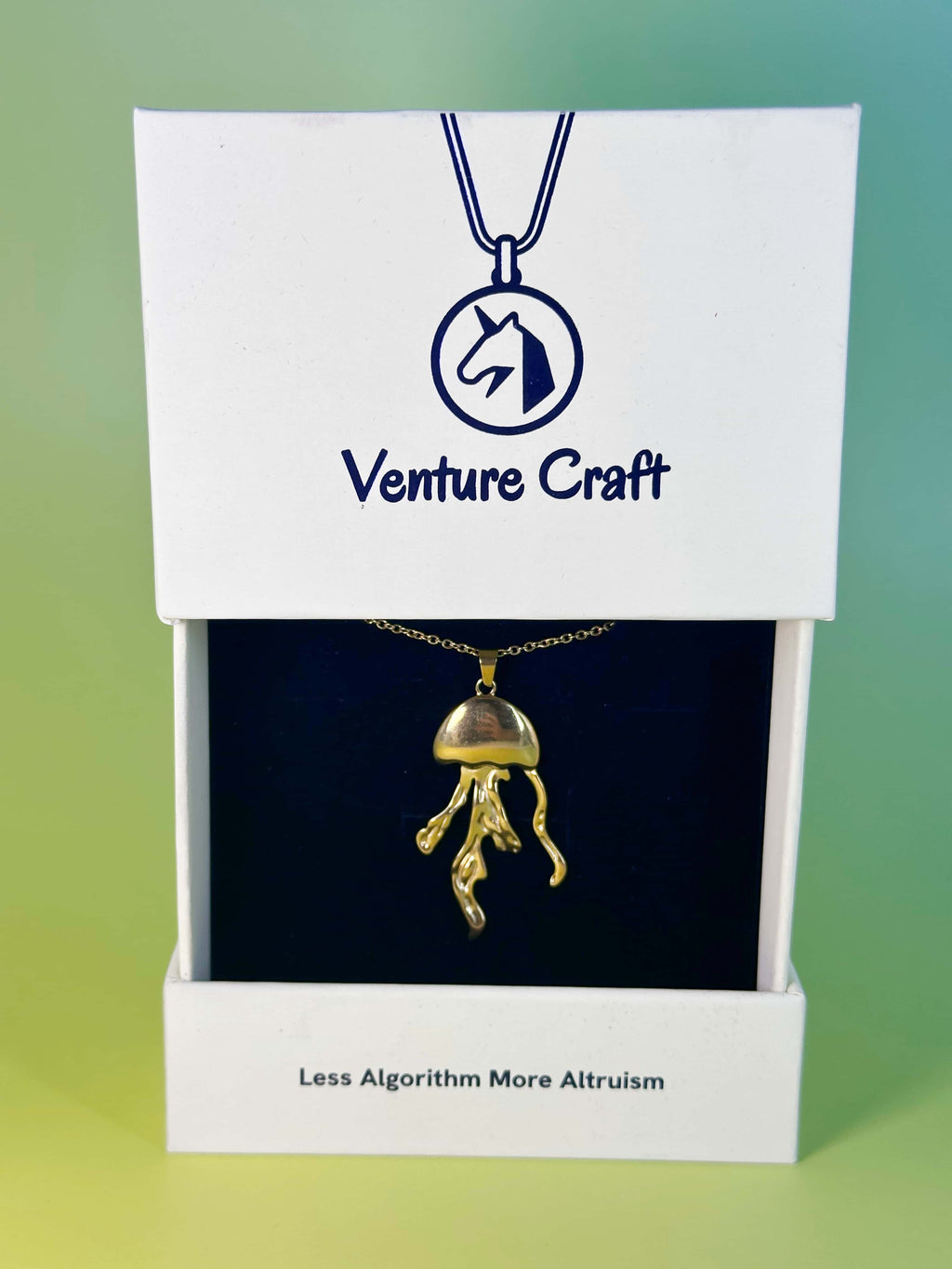 Premium gold jellyfish necklace presented inside its custom white Venture Craft gift box, featuring the brand's unicorn logo and the slogan 'Less Algorithm More Altruism'