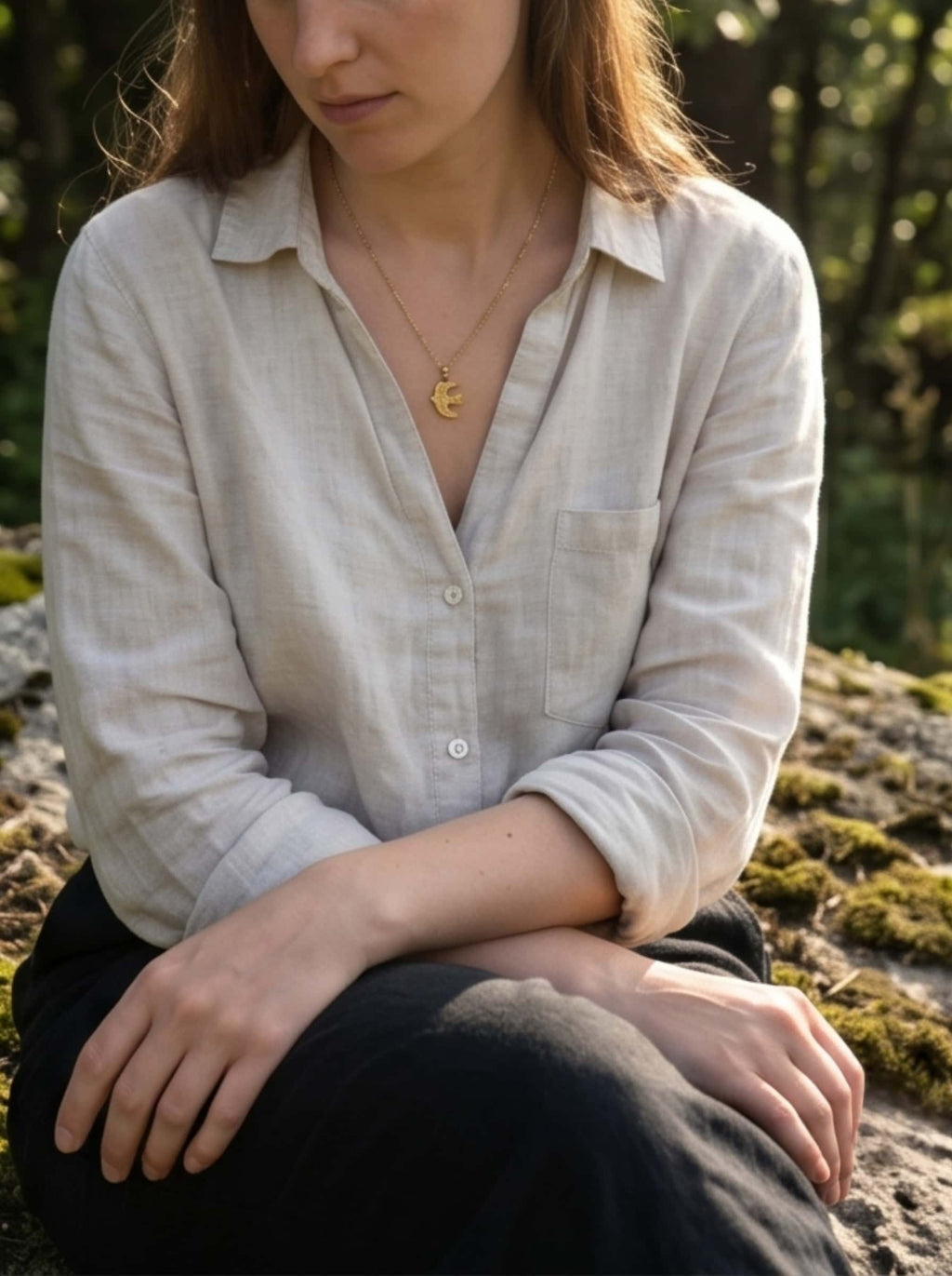 Lifestyle shot of a woman wearing a gold bird necklace while sitting in a peaceful forest setting with mossy rocks